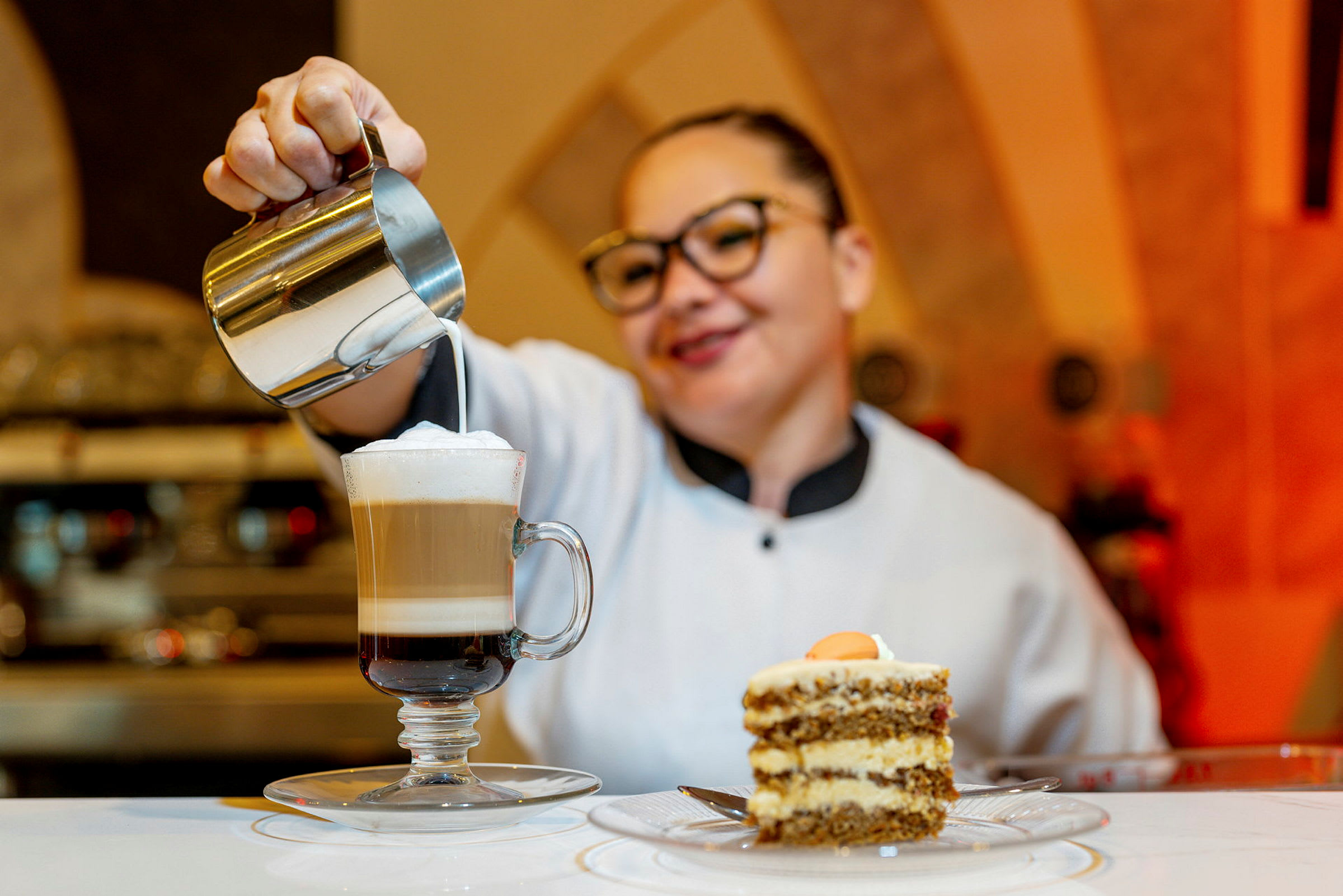 En kvinna häller mjölk i en kaffe latte framför en tårtbit på ett bord. (AI-genererad text)