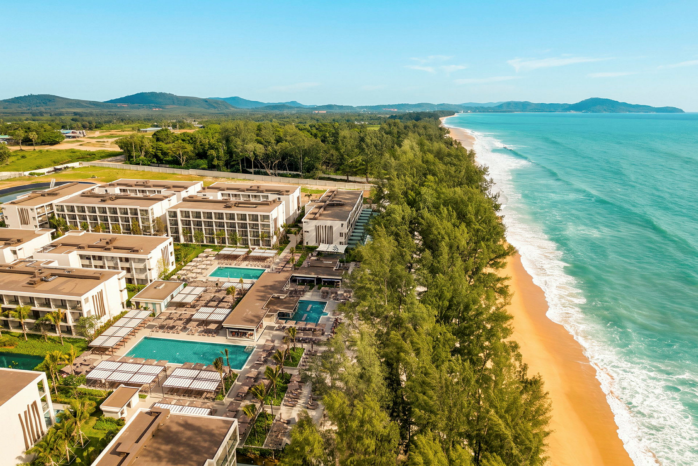 Flygfoto över ett modernt hotellkomplex med flera byggnader, pooler och trädgårdar, beläget vid en bred strand med gyllene sand och omgiven av gröna träd. (AI-genererad text)