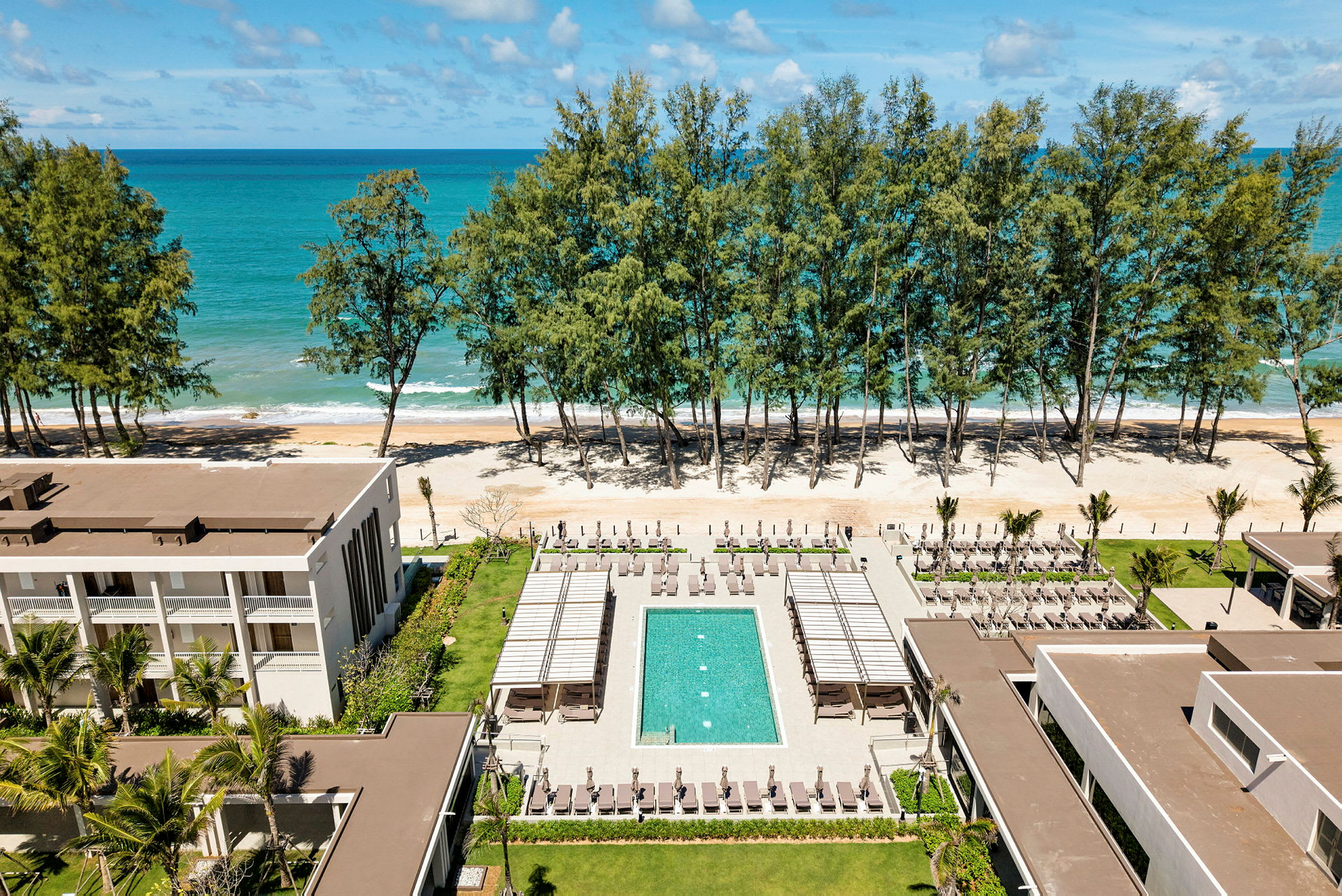 En hotellanläggning med swimmingpool, solstolar och grönområden vid strandkanten, med havet och träd i bakgrunden. (AI-genererad text)