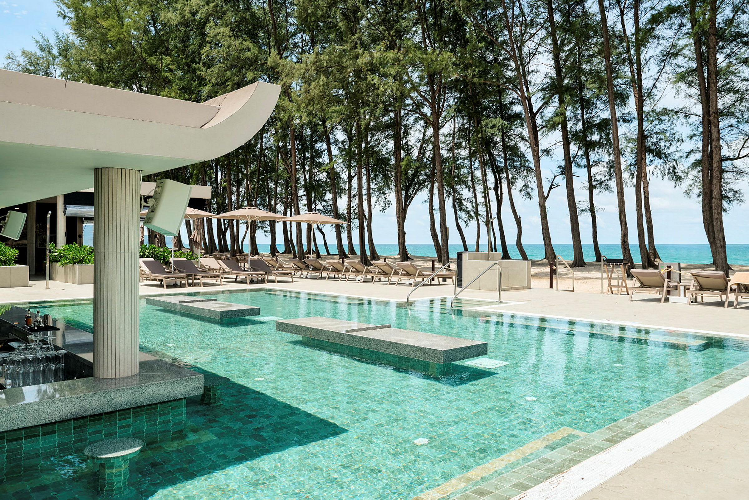 En lyxig pool med bar i förgrunden vid stranden, omgiven av solstolar och parasoller samt tallträd i bakgrunden. (AI-genererad text)