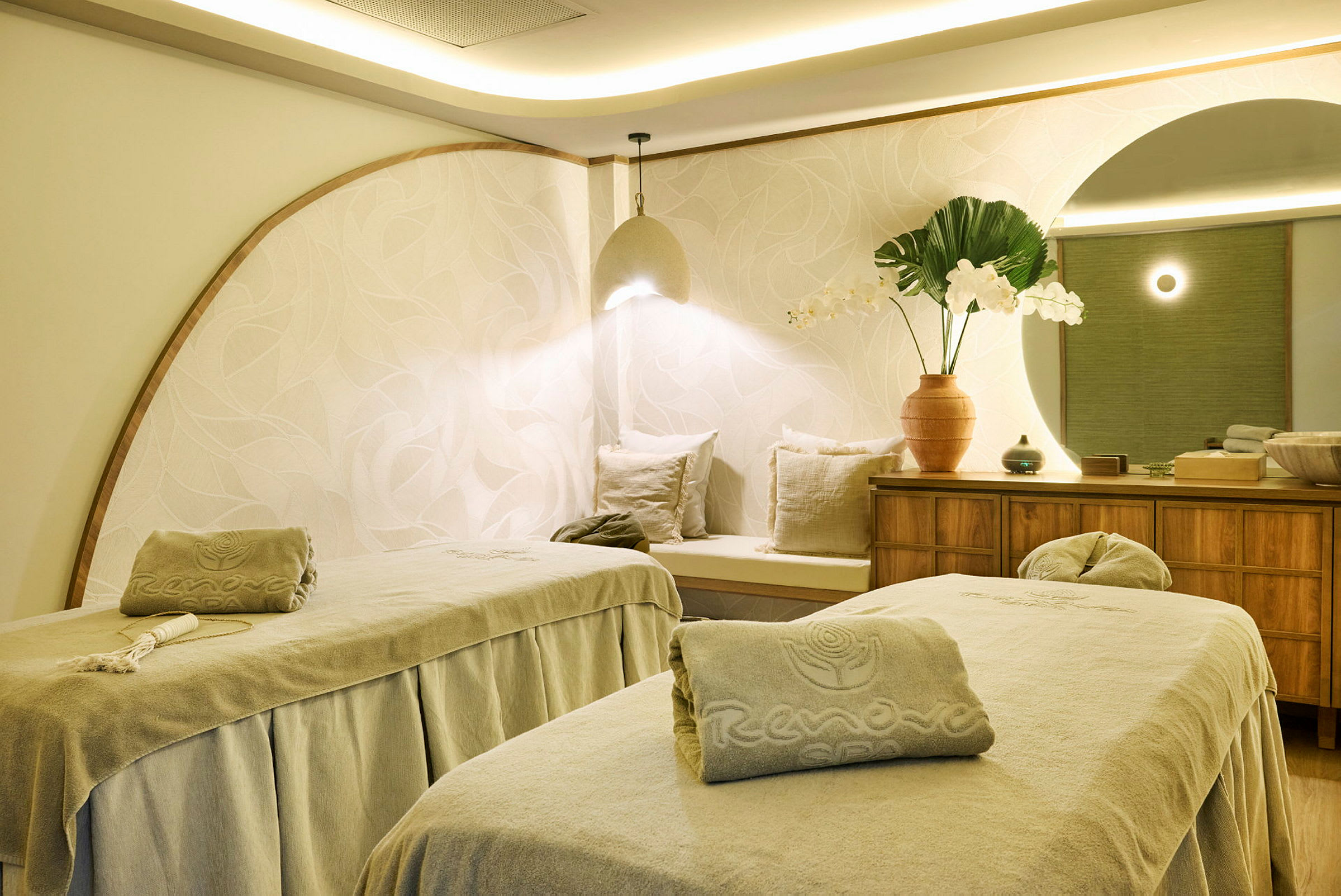 Två massagebänkar i ett väl upplyst och stilfullt inrett spa-rum. På en skänk står dekorativa element som en vas med blommor. (AI-genererad text)