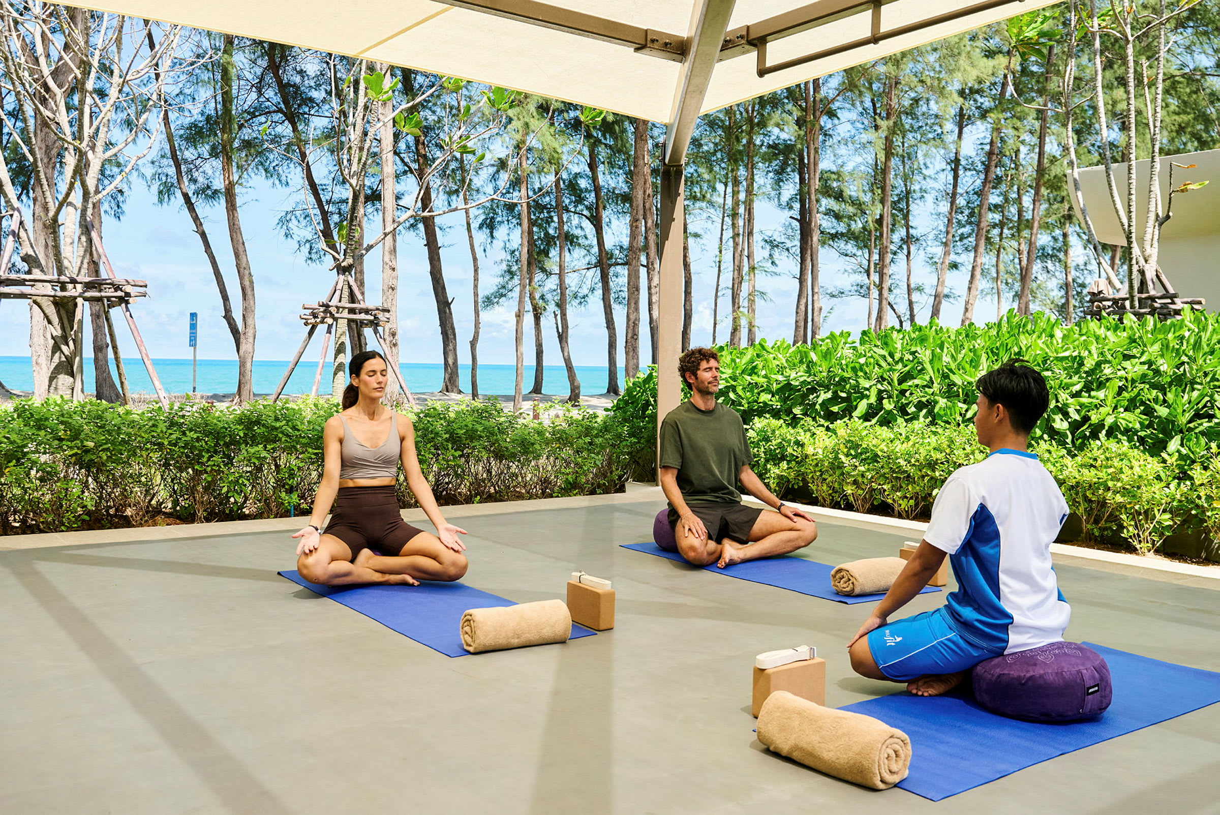 Tre personer sitter på yogamattor utomhus och mediterar. Bakom dem finns gröna buskar, träd och en strand med havet i bakgrunden. (AI-genererad text)