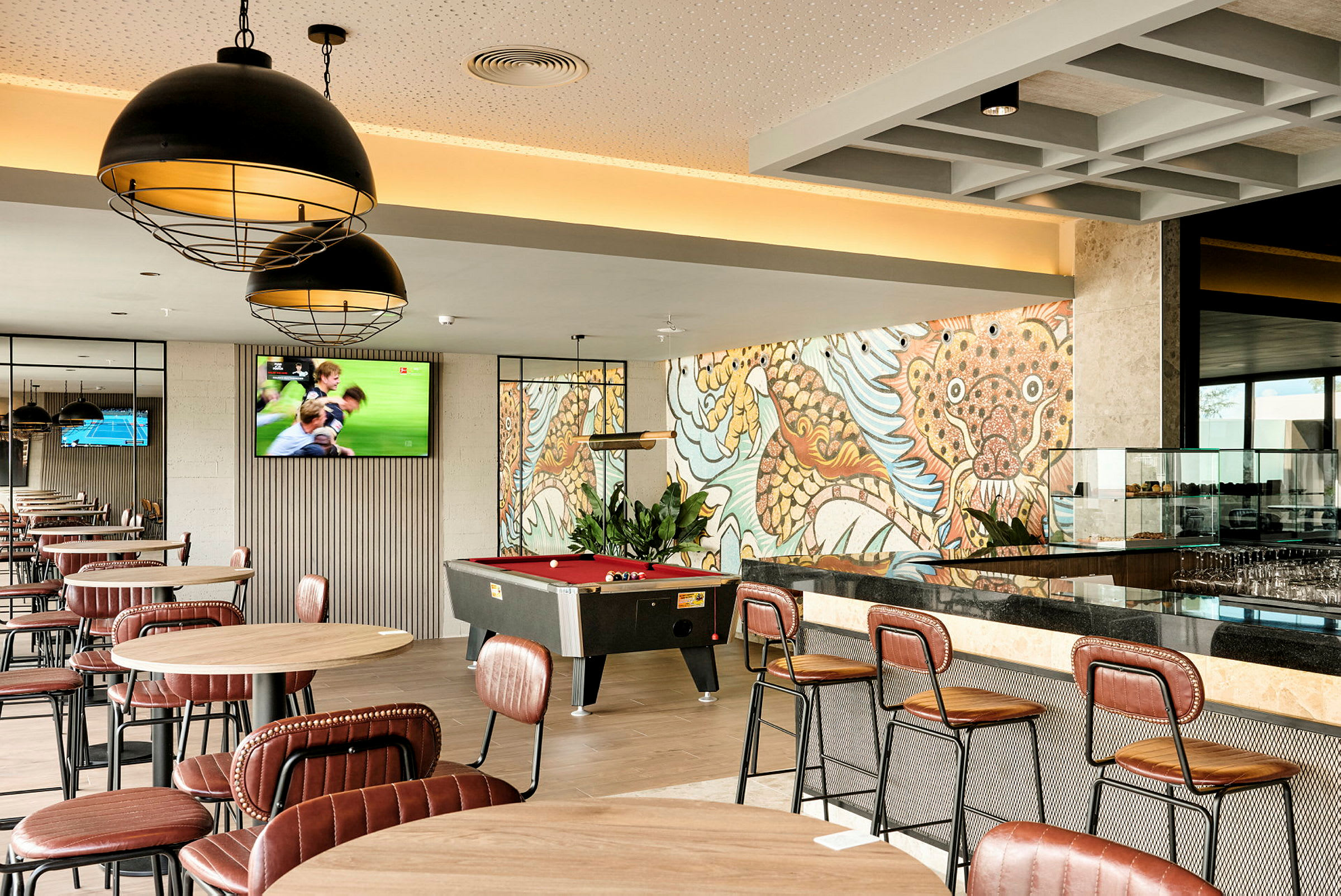 Ett café eller loungemiljö med bord och stolar, en biljardbord och en färgrik väggmålning i bakgrunden. (AI-genererad text)