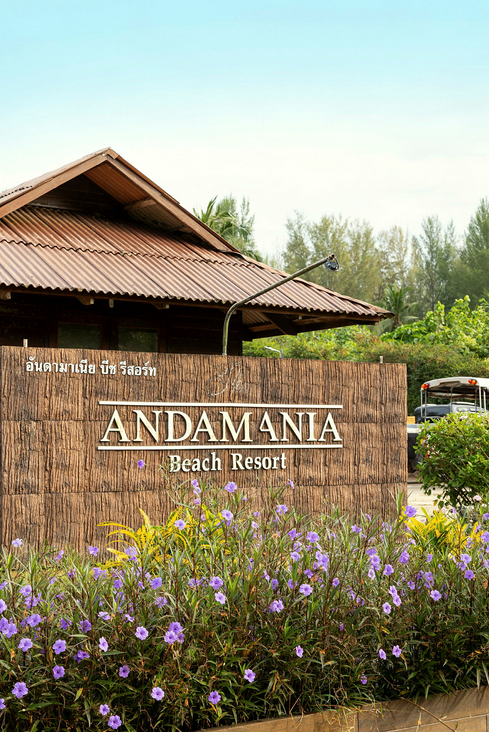 Entrén till Andamania Beach Resort, med en skylt omgiven av lila blommor och en byggnad med ett plåttak i bakgrunden. (AI-genererad text)