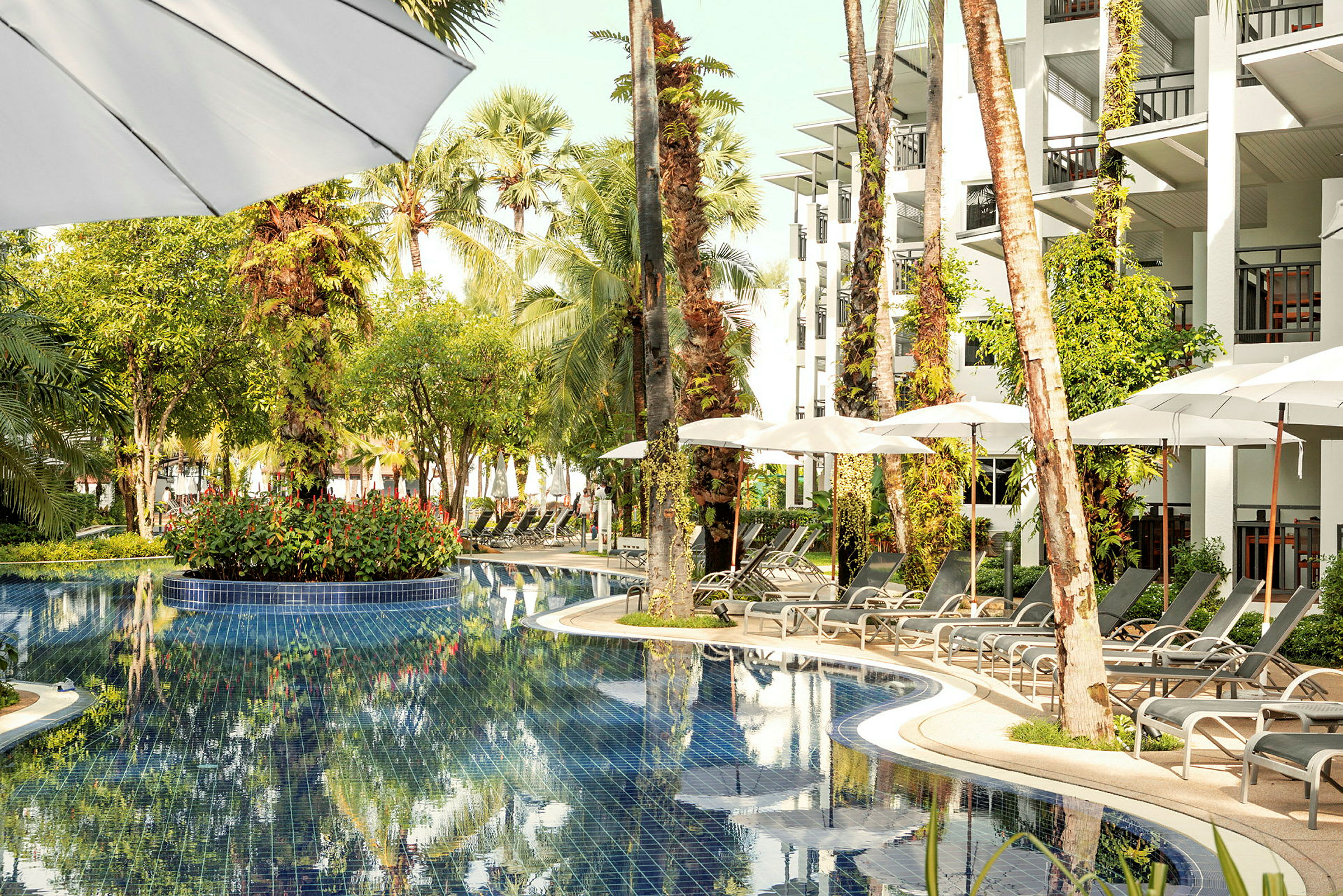 Bild av en pool omgiven av solstolar, parasoller och grönska vid hotellbyggnad. (AI-genererad text)