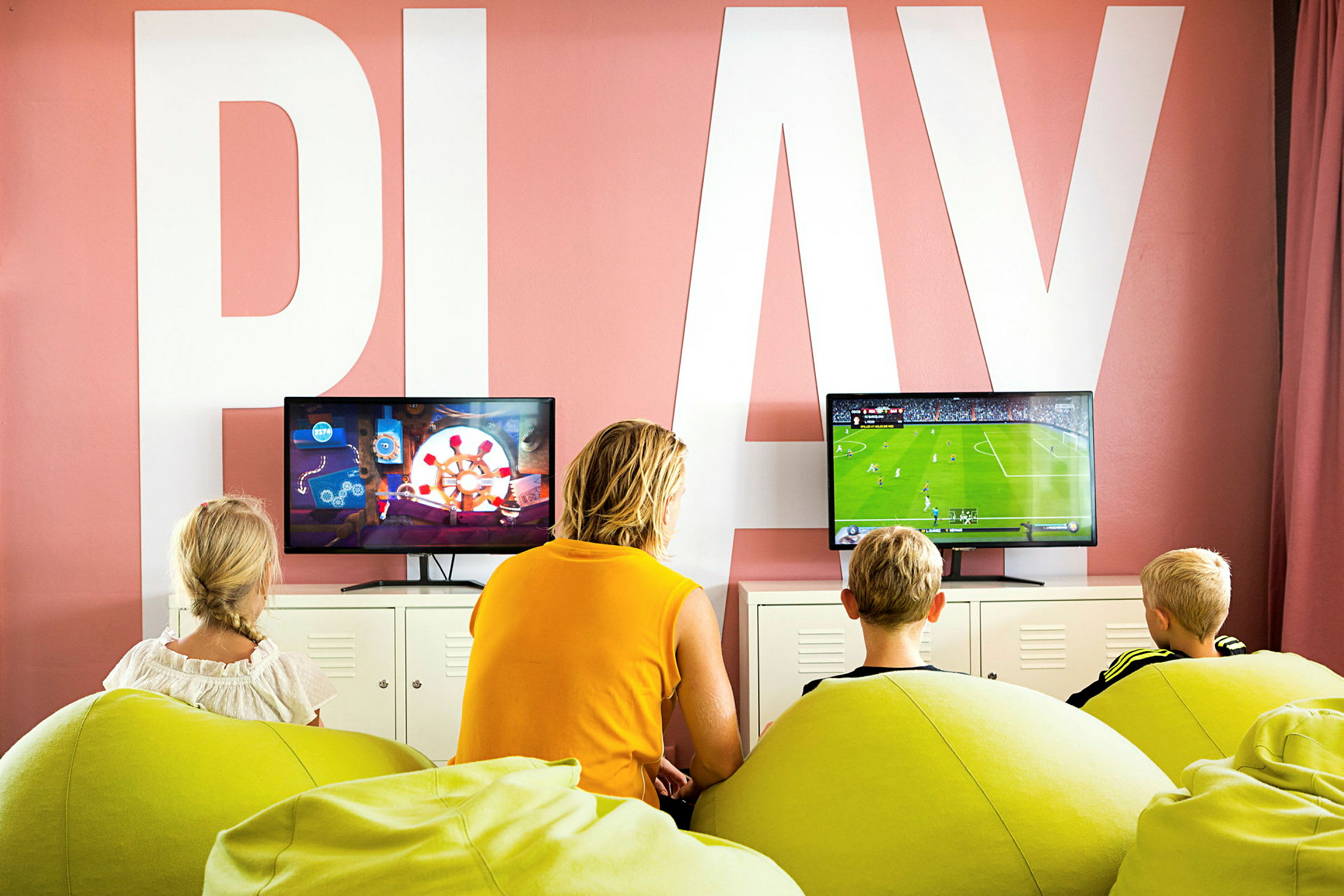 Tre barn och en vuxen sitter på gröna säckstolar och spelar TV-spel framför två skärmar i ett rum med texten 'PLAY' på väggen. (AI-genererad text)