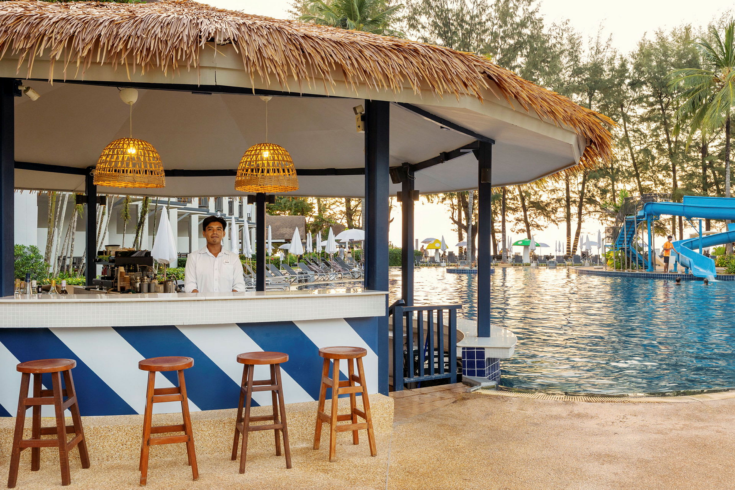 En strandbar med halmtak vid en pool. Det finns barstolar framför baren och omgivningen inkluderar solstolar och parasoller. (AI-genererad text)
