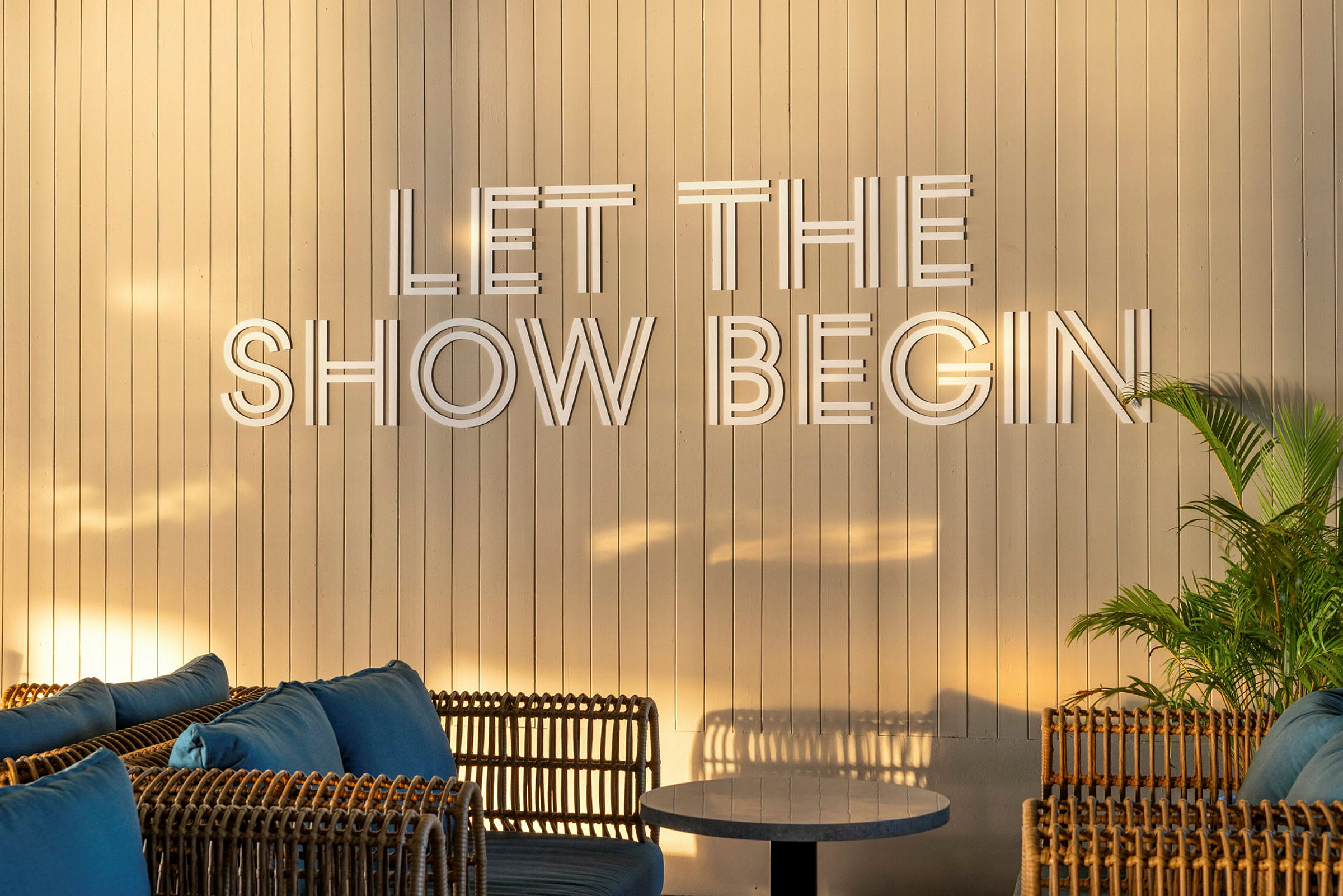 En ljus träpanelvägg med texten "Let the show begin", omgiven av rottingmöbler och gröna växter. (AI-genererad text)
