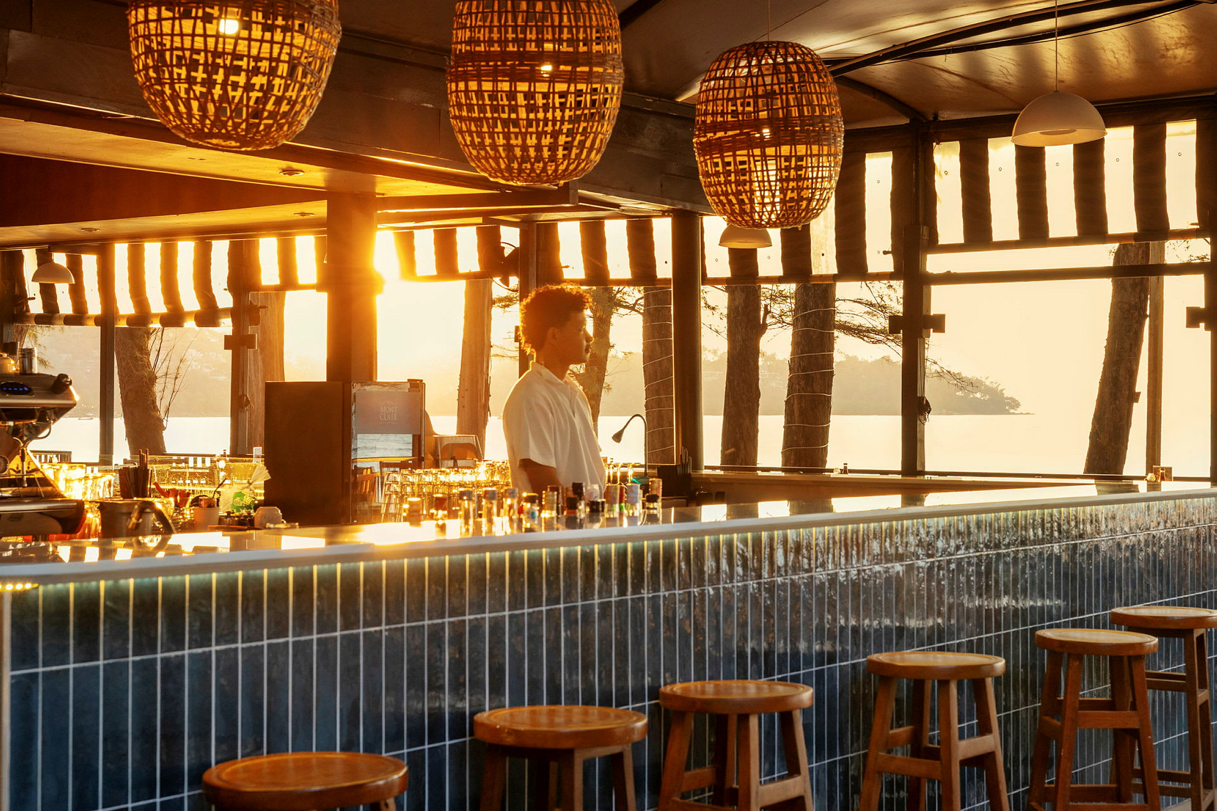 En stilfull bar belyst av varmt solljus genom fönstren, med runda träpallar framför en lång bardisk. (AI-genererad text)