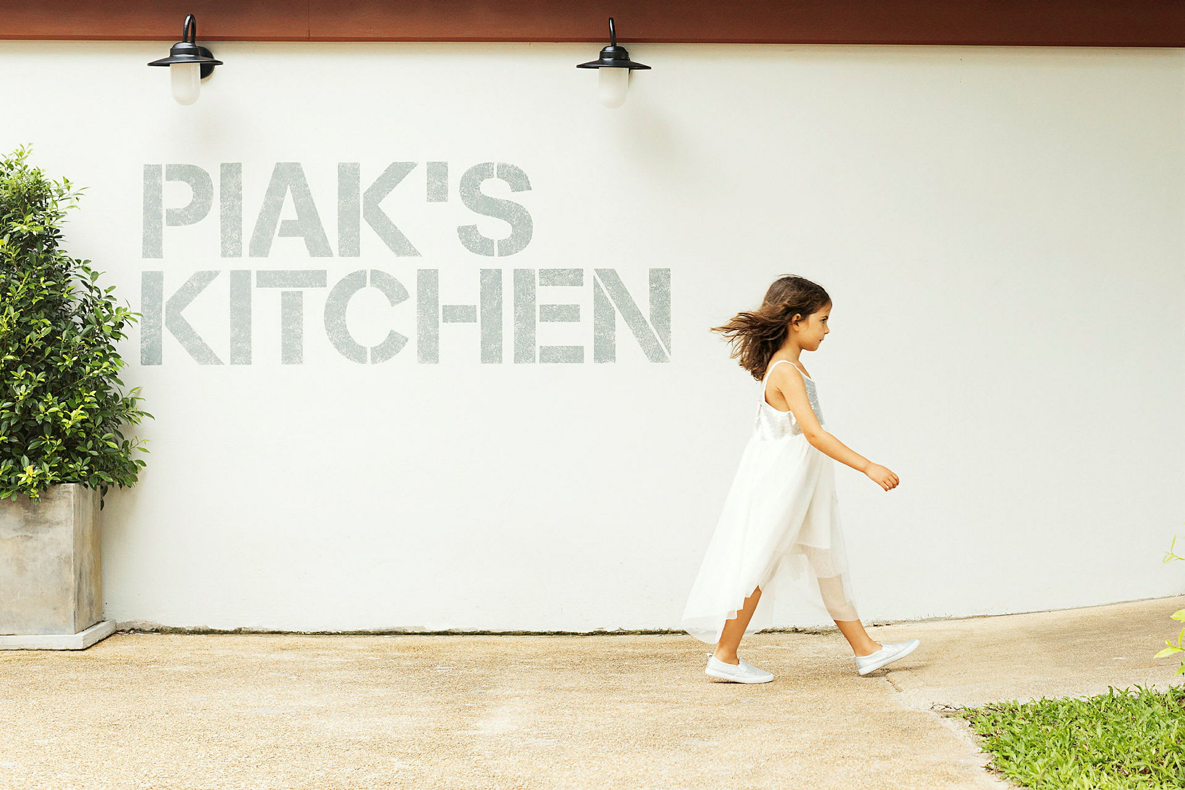 En person går förbi en vit vägg med texten 'PIAK'S KITCHEN' och en lampa samt en krukväxt synliga. (AI-genererad text)
