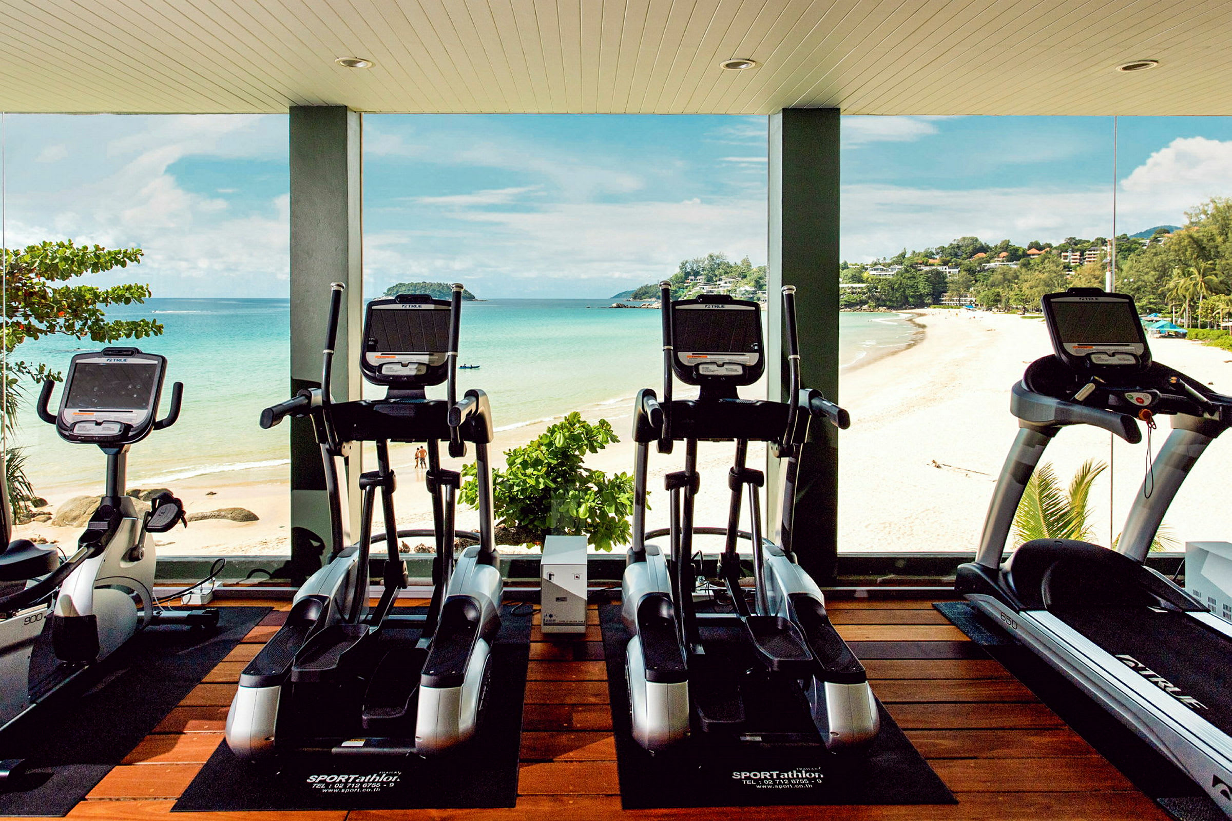 Bild av ett gym med motionscyklar och löpband framför stora fönster med utsikt över en strand och hav. (AI-genererad text)