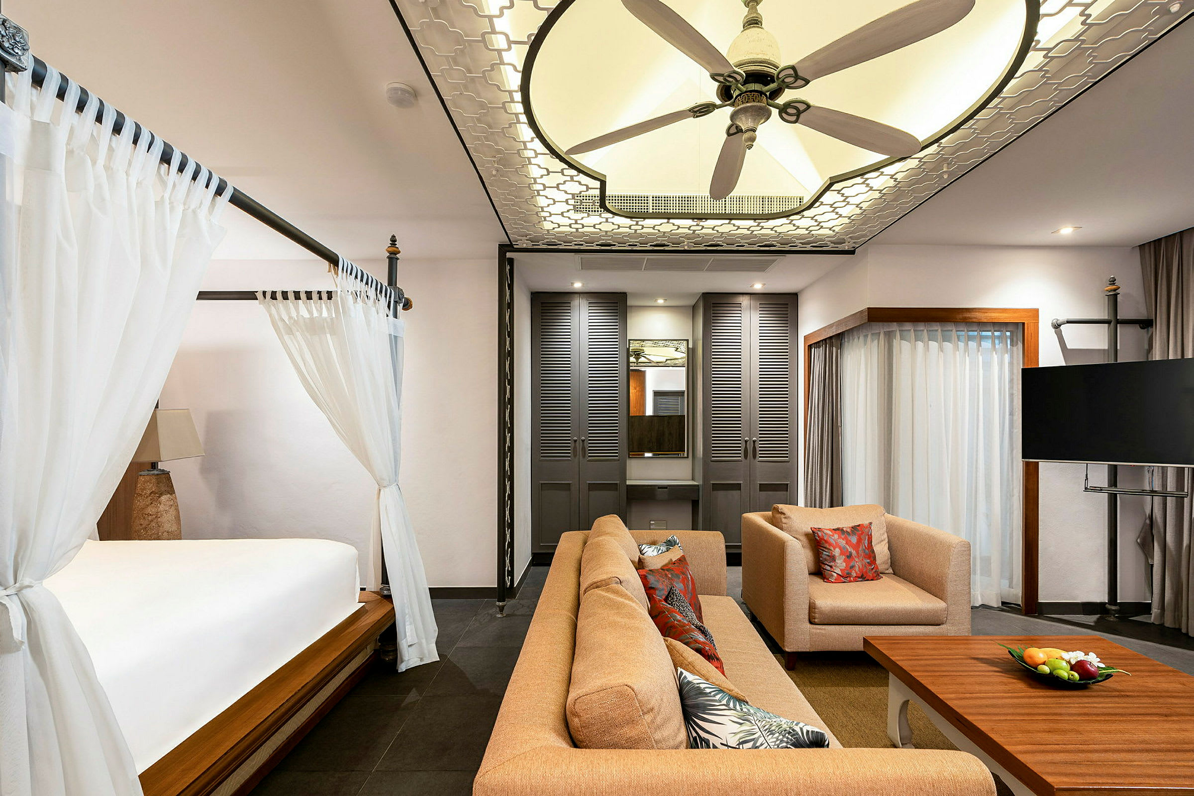 Ett elegant hotellrum med en säng med himmel, en sittgrupp med soffa och fåtölj, ett soffbord med fruktskål, en TV och en takfläkt. (AI-genererad text)