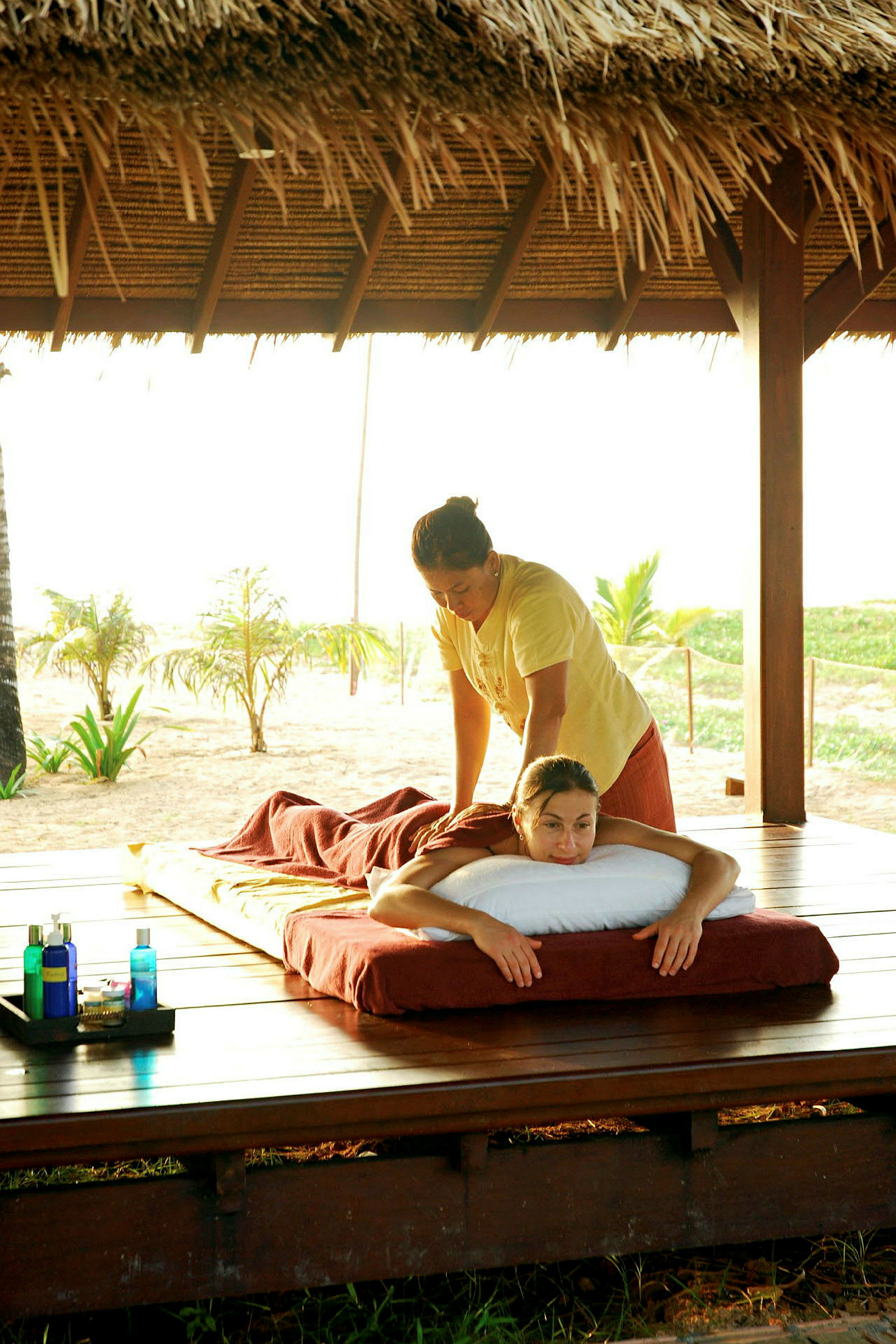 En person får massage på en utomhusterrass med strand och palmträd i bakgrunden. På sidan finns flaskor med massageolja. (AI-genererad text)