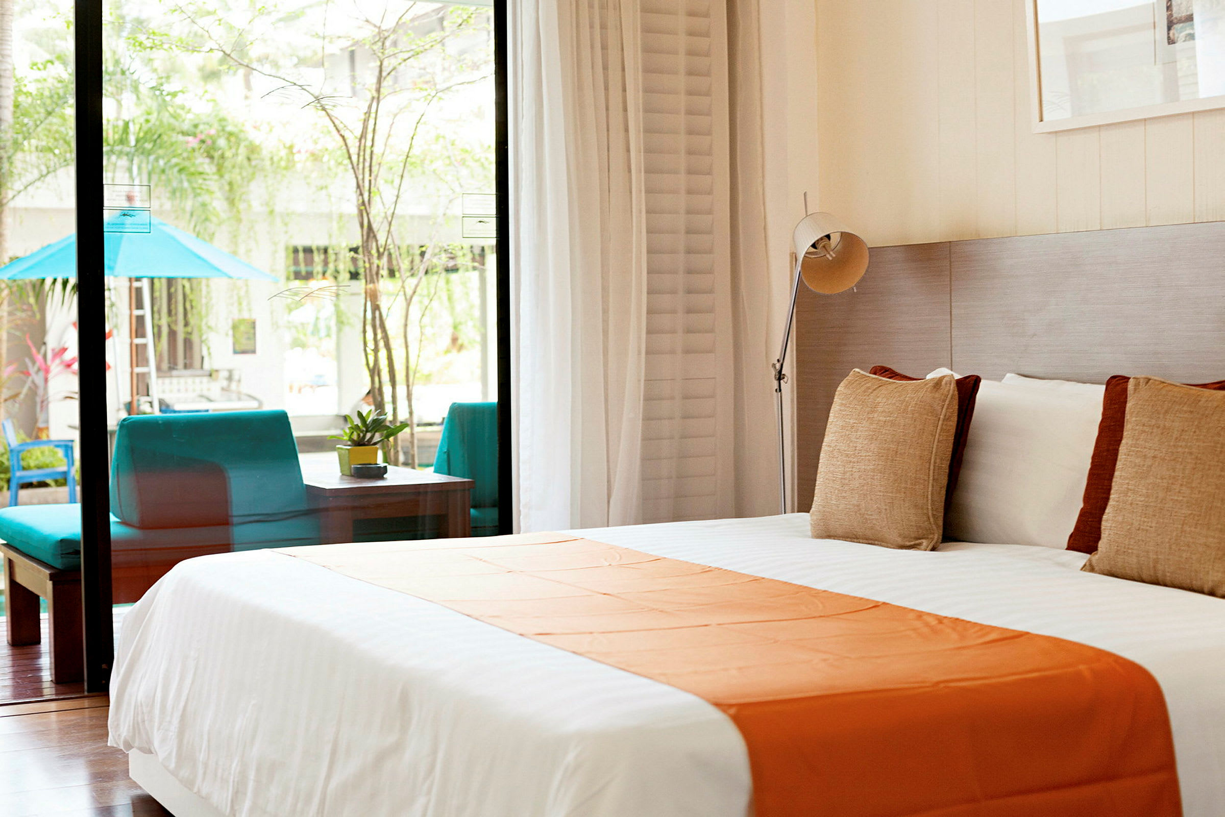 Bild av ett ljust hotellrum med en stor säng klädd i vitt och orange, stora fönster som vetter mot en uteplats med sittgrupp och grönska. (AI-genererad text)