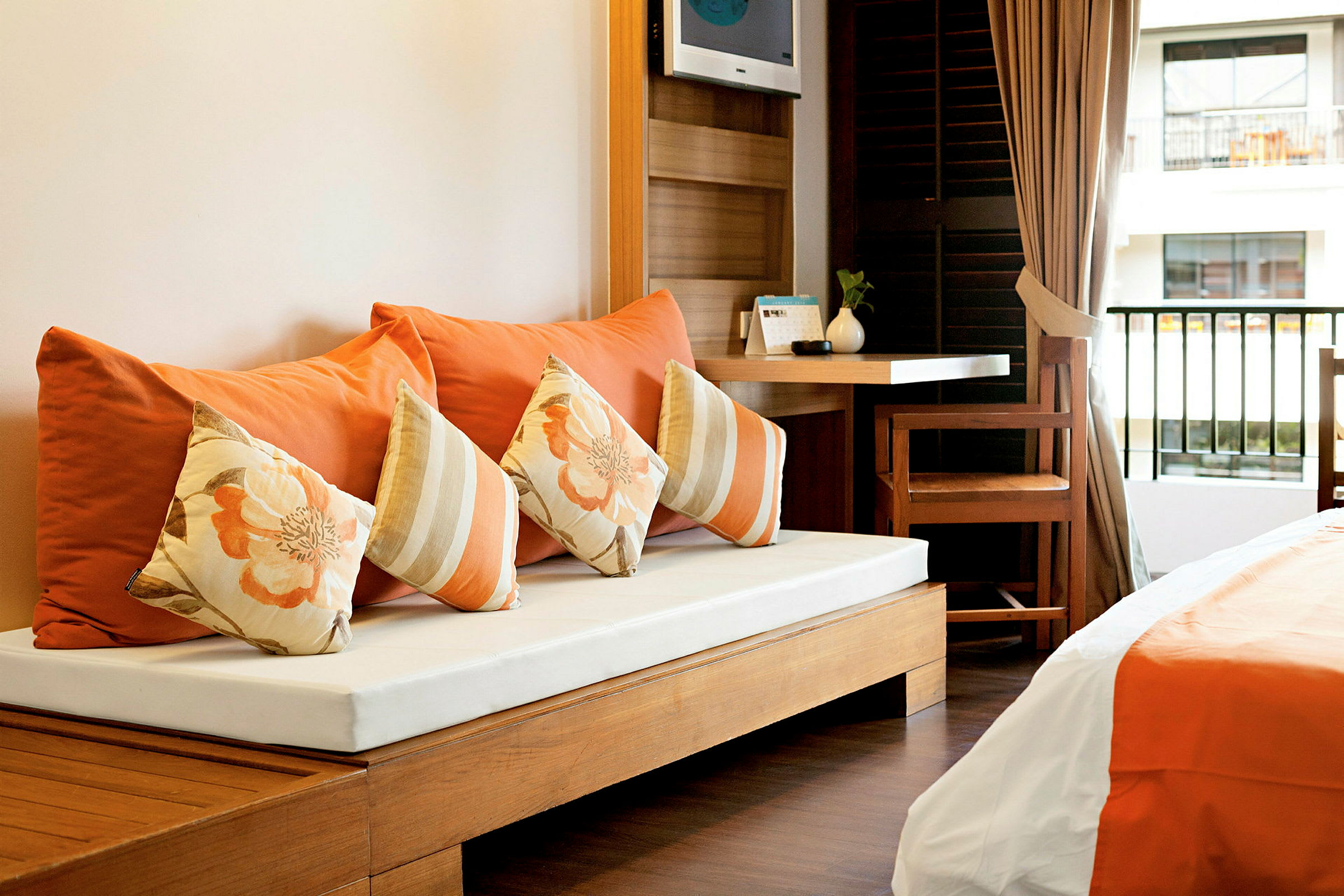 Ett hotellrum med en träbänk, dekorerade kuddar i orange och blommigt mönster, skrivbord, stol och säng med orange sängkläder. (AI-genererad text)