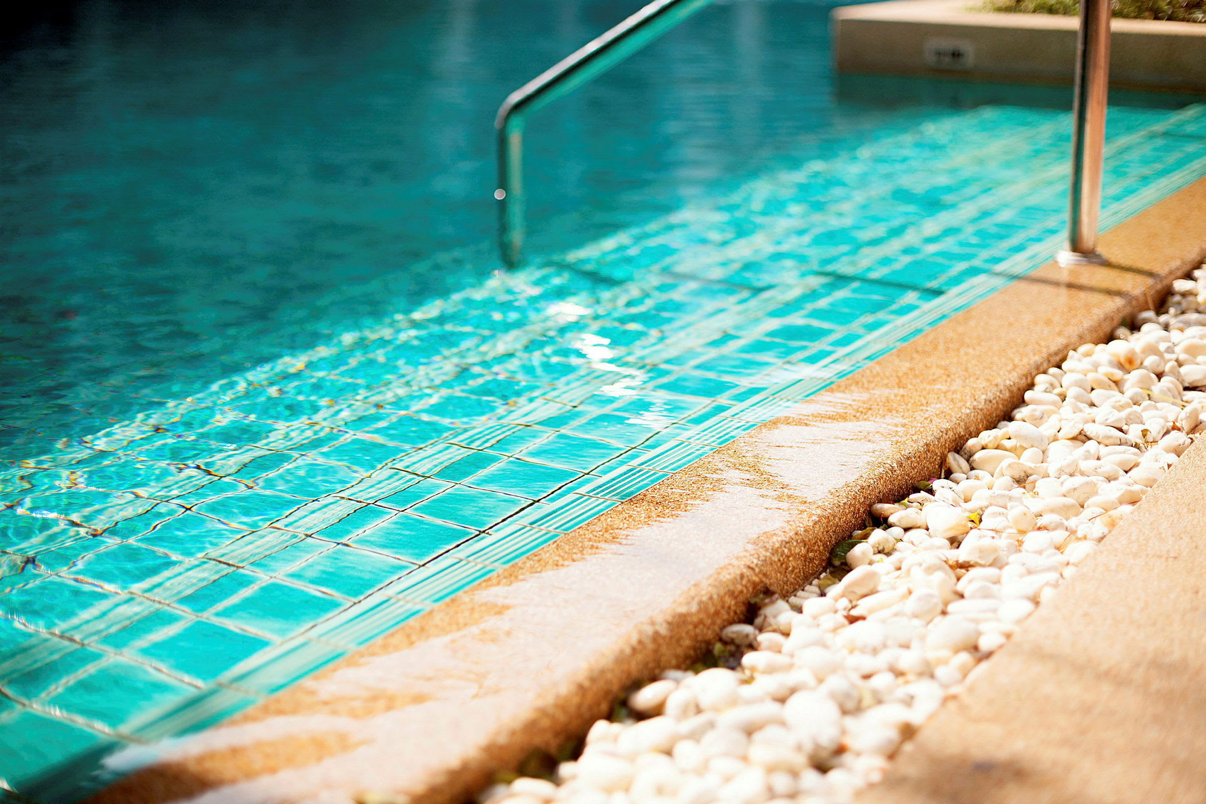 Närbild av kanten på en swimmingpool med blått kakel. Detaljer av vattenyta, en metallstång samt vita prydnadsstenar är synliga. (AI-genererad text)