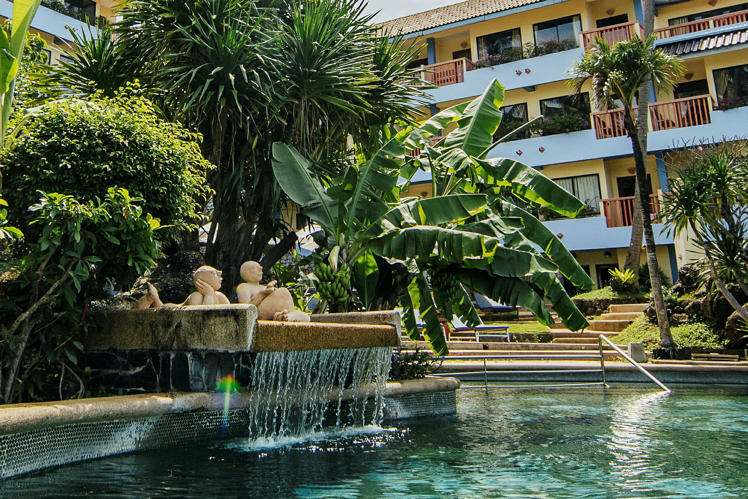 En pool med vattenfall och närliggande vegetation framför hotell byggnad. (AI-genererad text)