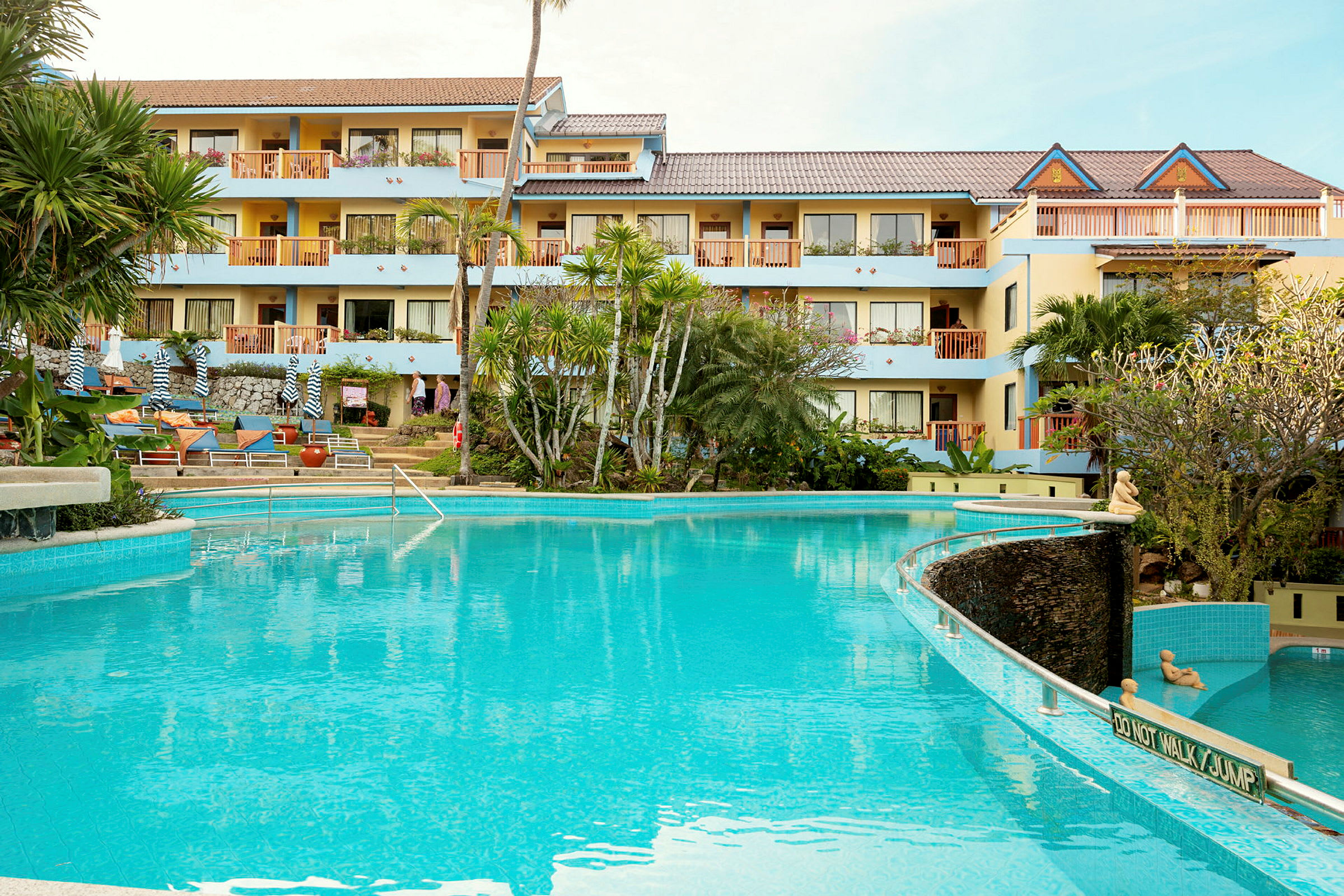 Hotellbyggnad med flera våningar synlig bakom en klarblå swimmingpool, omgiven av tropiska växter. (AI-genererad text)