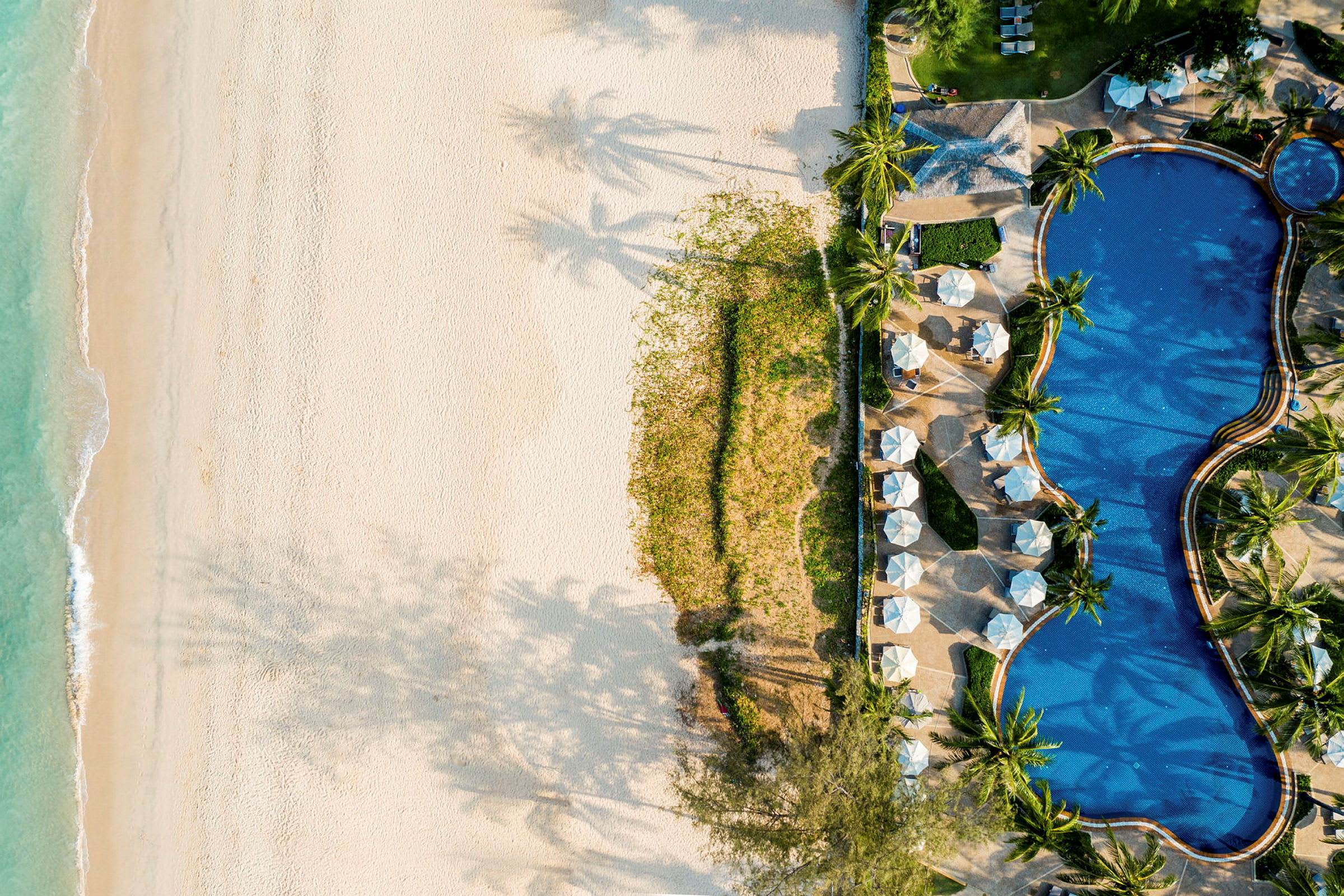 Flygfoto av en strand med sand och grönt nära en pool med parasoller. (AI-genererad text)