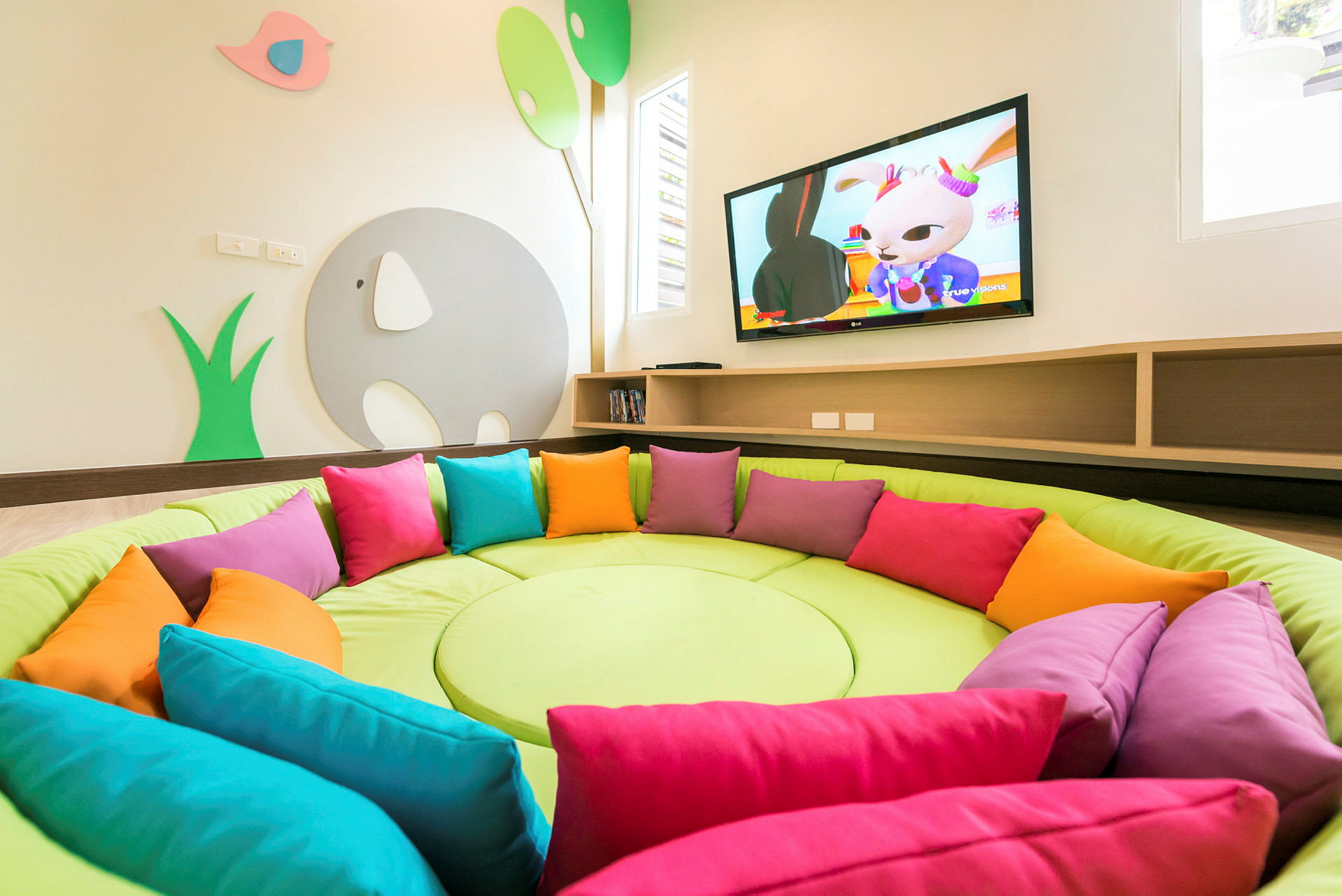 En rund sittplats med färgglada kuddar framför en vägg med TV och dekorativt barnmotiv. (AI-genererad text)
