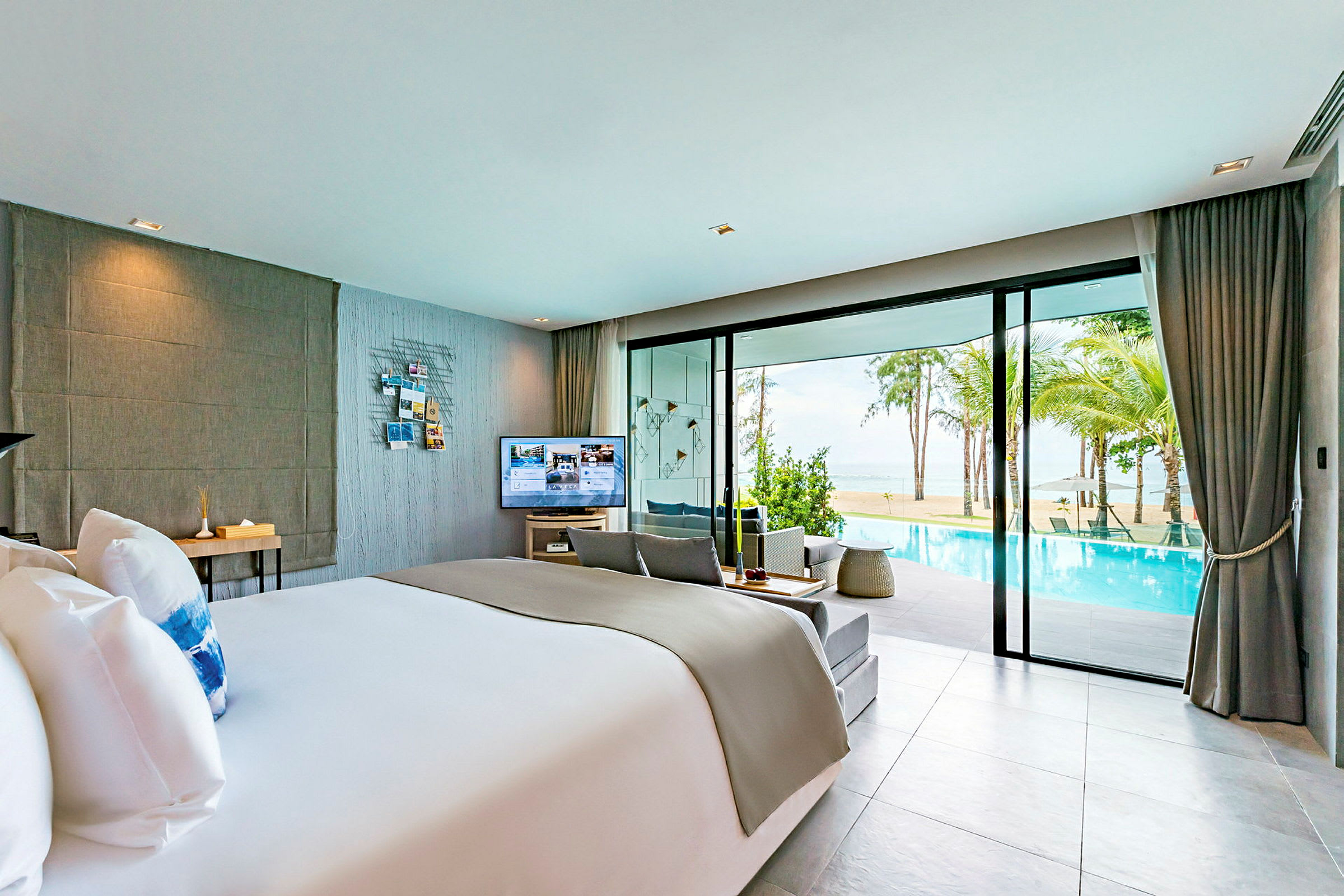 Ett modernt hotellrum med en stor säng, sittmöbler och en TV. Glasväggar visar utsikt över en pool, palmer och havet. (AI-genererad text)