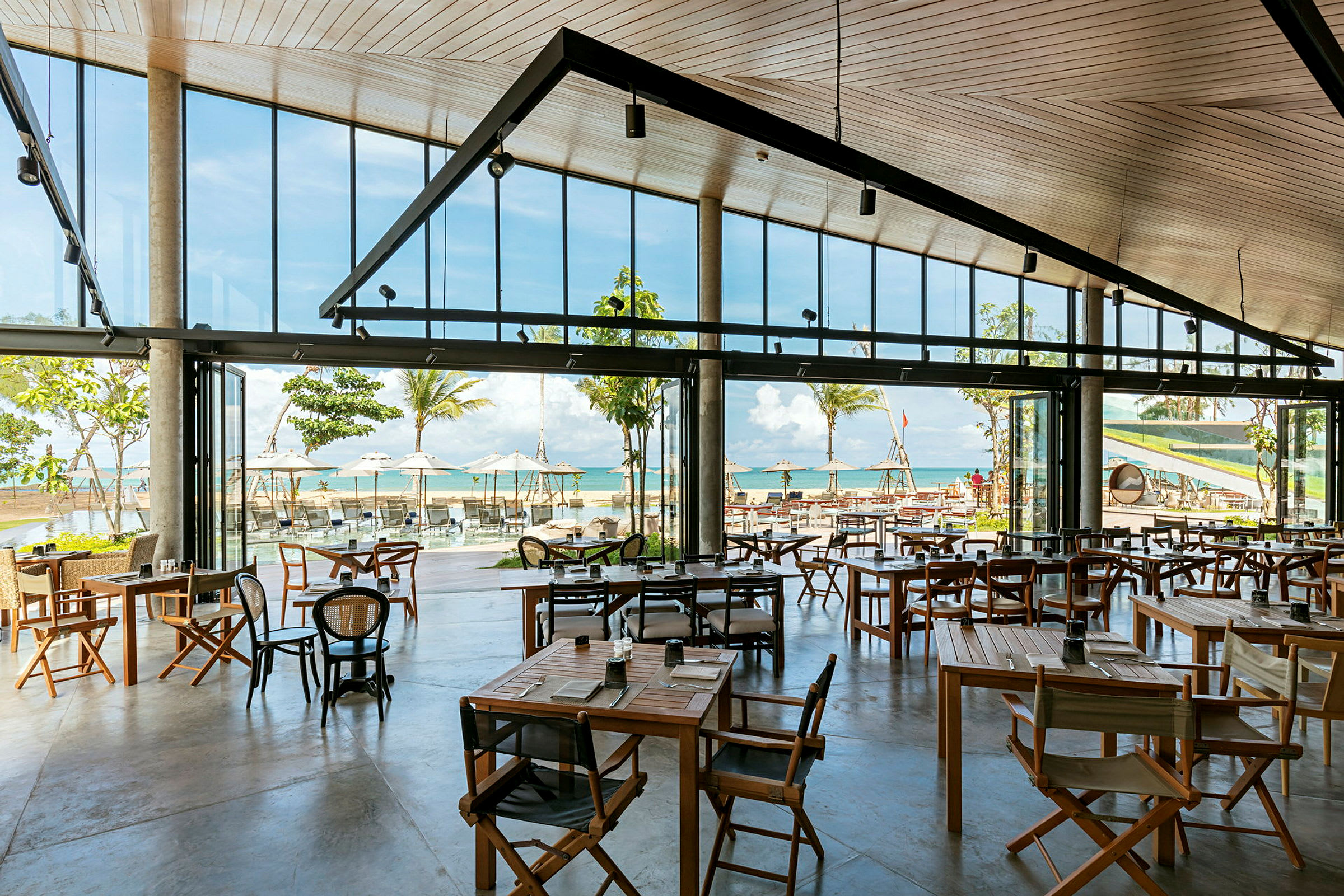 Ett öppet och ljust restaurangområde med stora fönster som vetter mot stranden och havet med palmer och parasoller. (AI-genererad text)