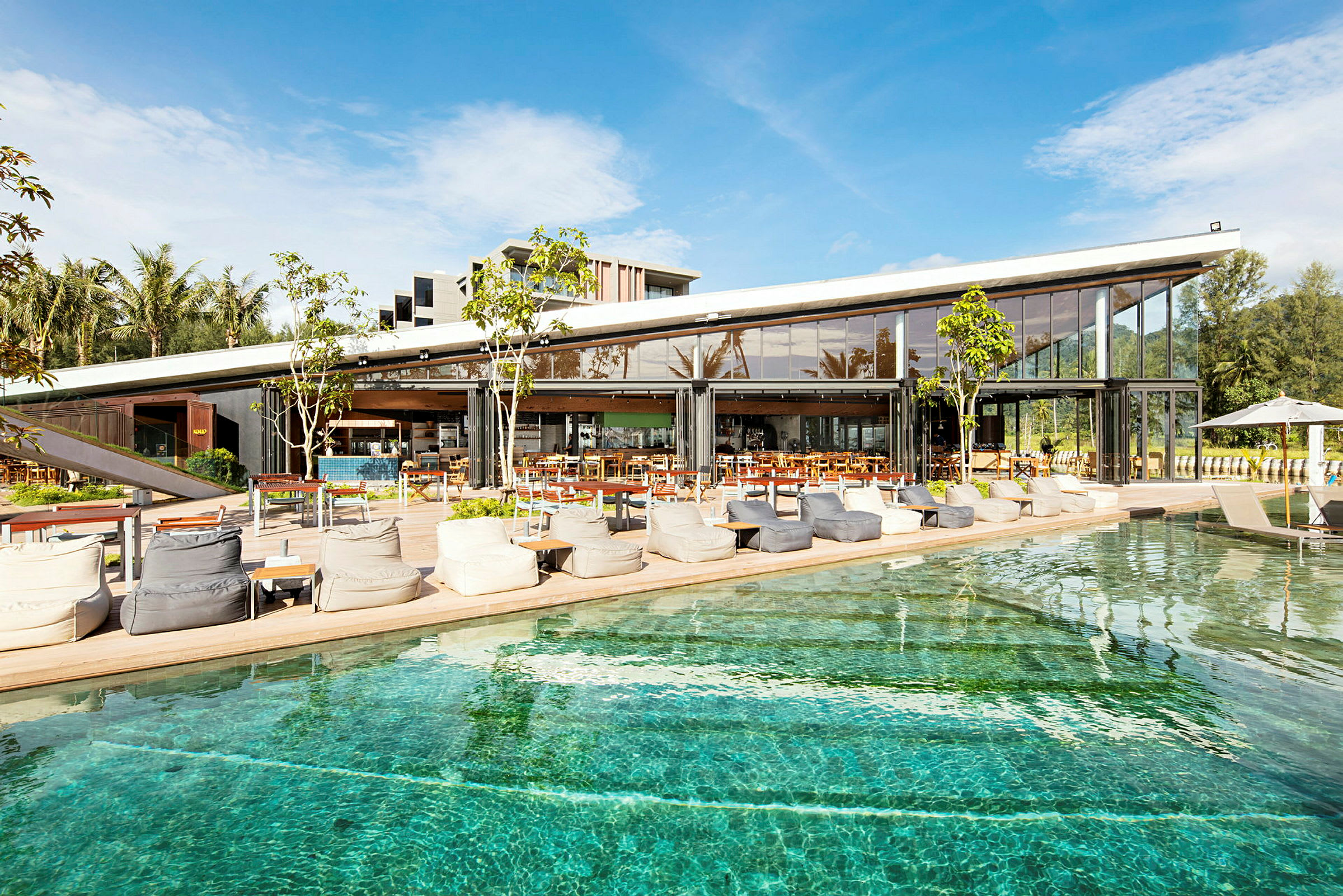 Ett modernt café eller restaurang med stora glasväggar ligger vid en pool under en klarblå himmel. (AI-genererad text)
