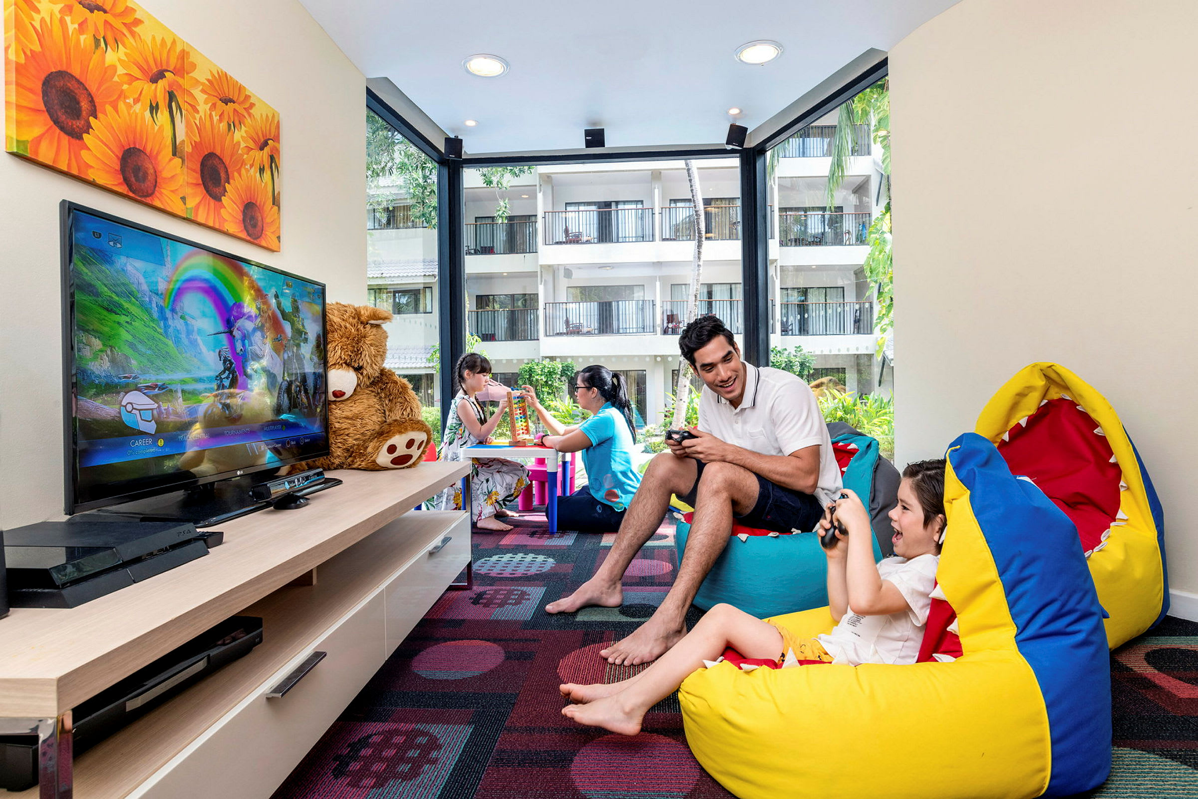 Ett rum med färgglatt inredning och beanbags, där barn och en vuxen spelar videospel och två personer bygger med klossar vid ett bord. (AI-genererad text)