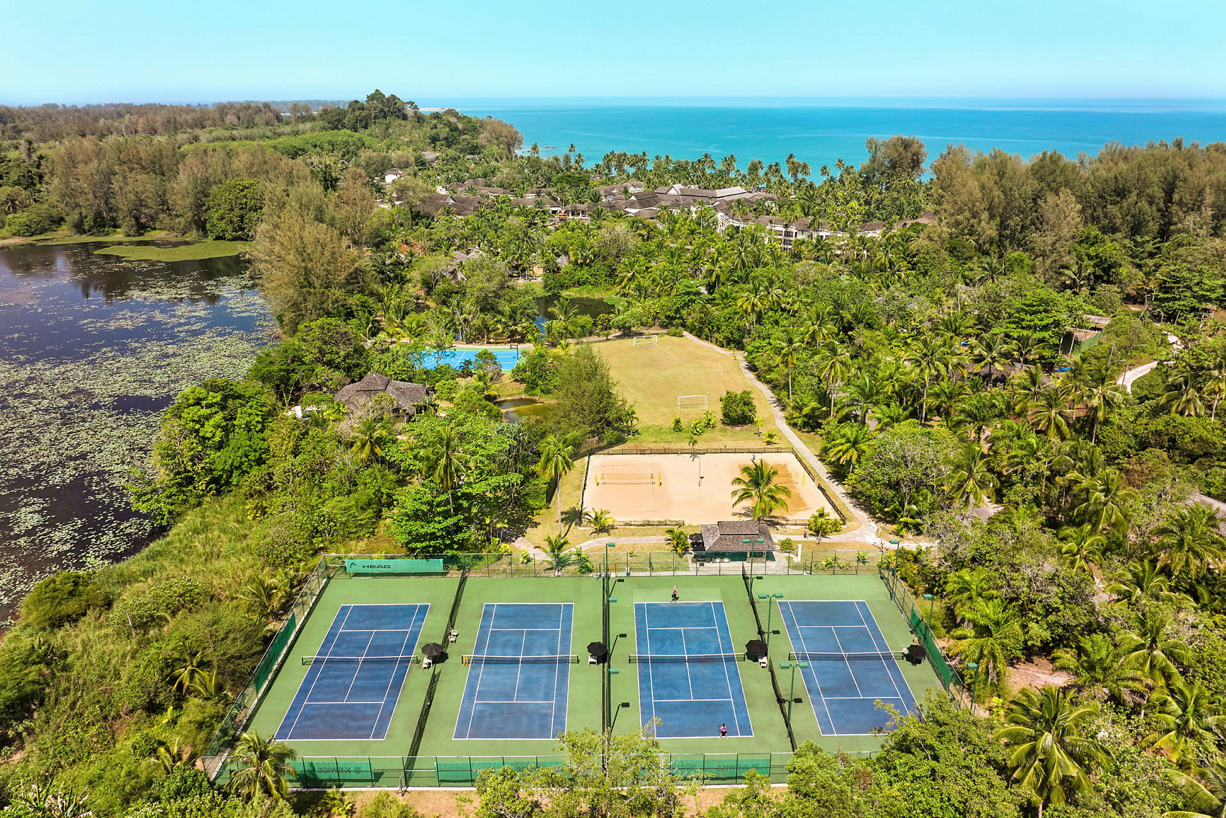 Flygfoto över ett grönskande landskap med tennisbanor, ett fotbollsplan och en sandplan. (AI-genererad text)
