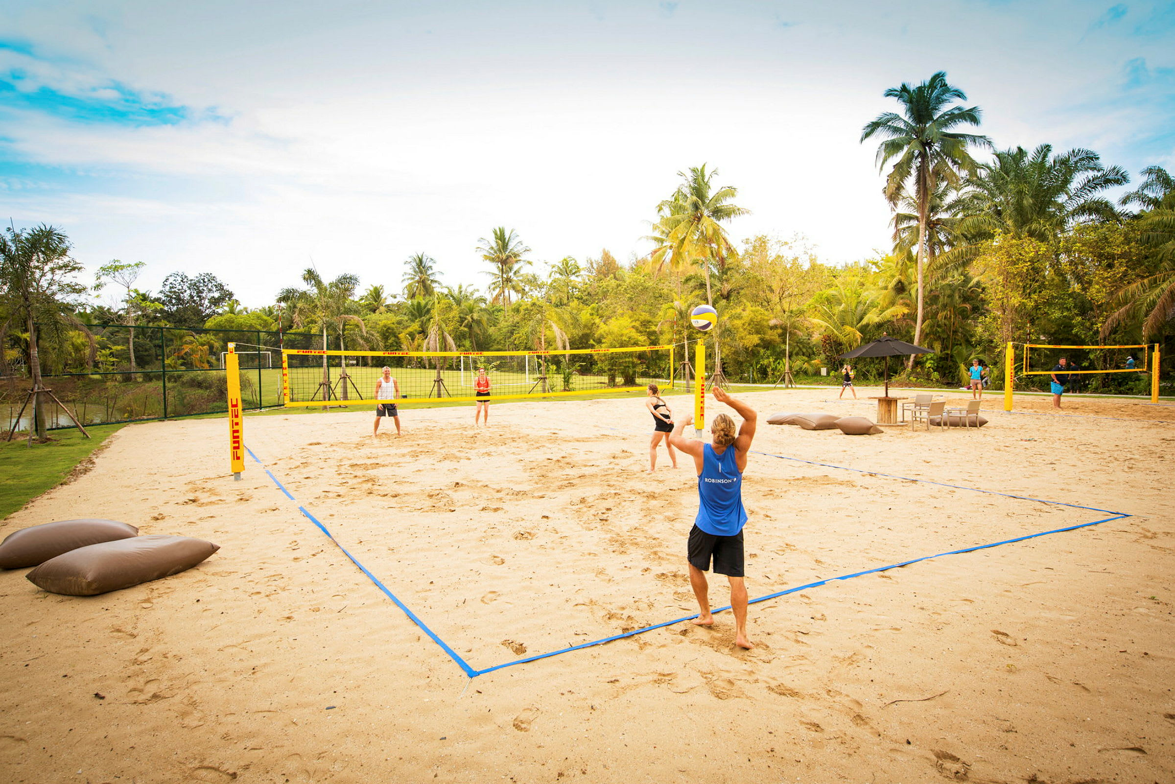 Flera personer spelar beachvolleyboll på en sandplan omgiven av palmer och grönska. (AI-genererad text)