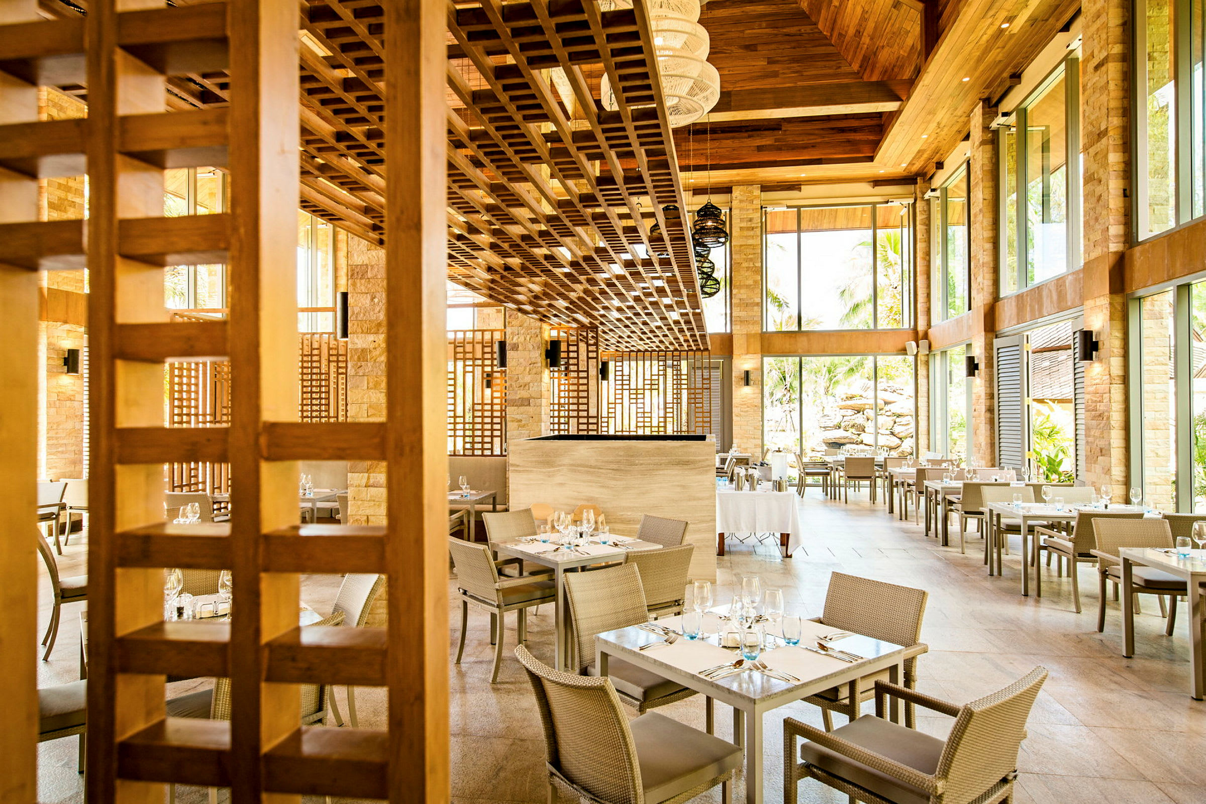 En ljus restaurang med sittplatser vid fyrkantiga bord och stora glasväggar som släpper in naturligt ljus. Taket är gjort av trä. (AI-genererad text)