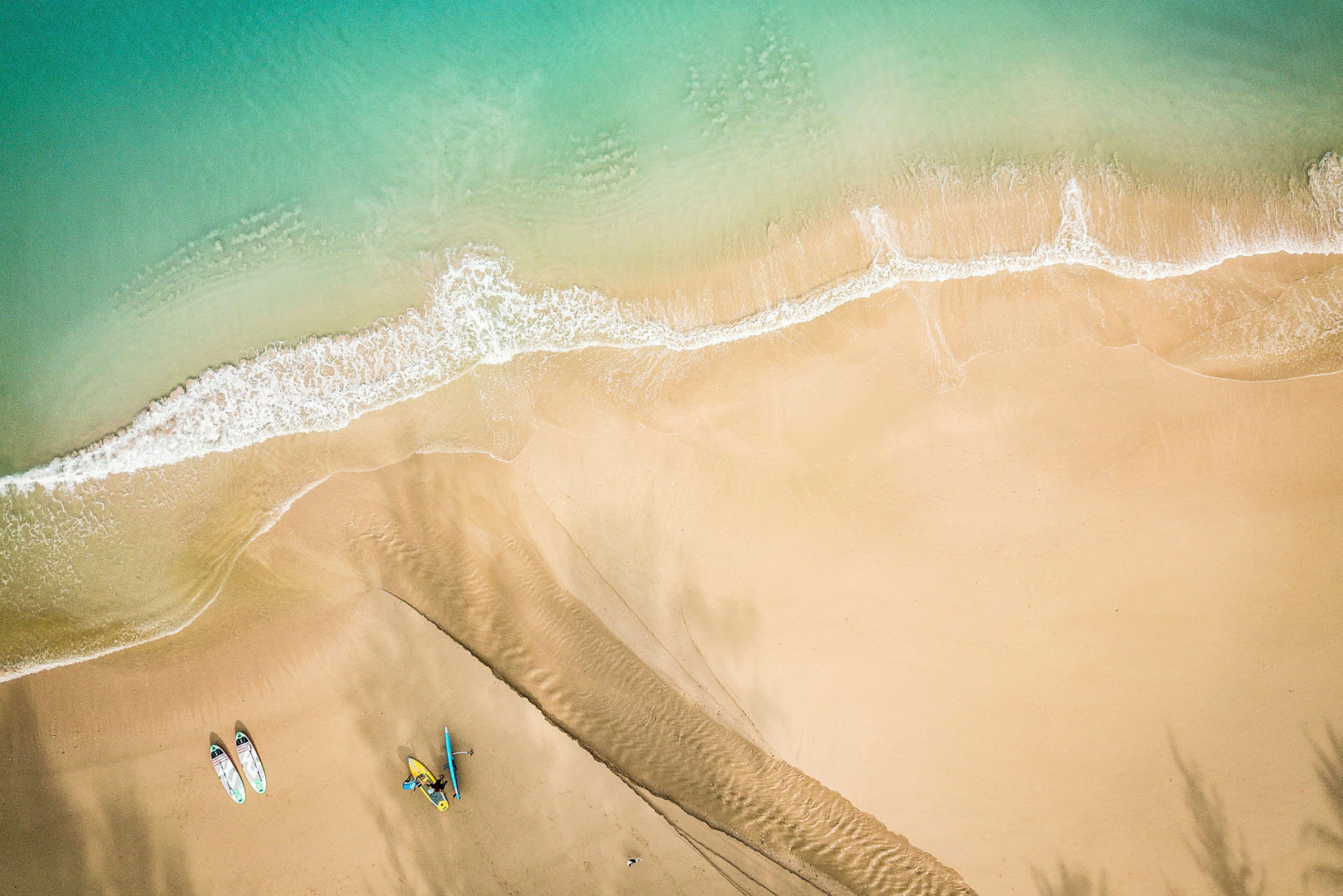 Flygfotografi av en sandstrand där vågor möter stranden. Två paddleboards och en kajak syns på sanden. (AI-genererad text)