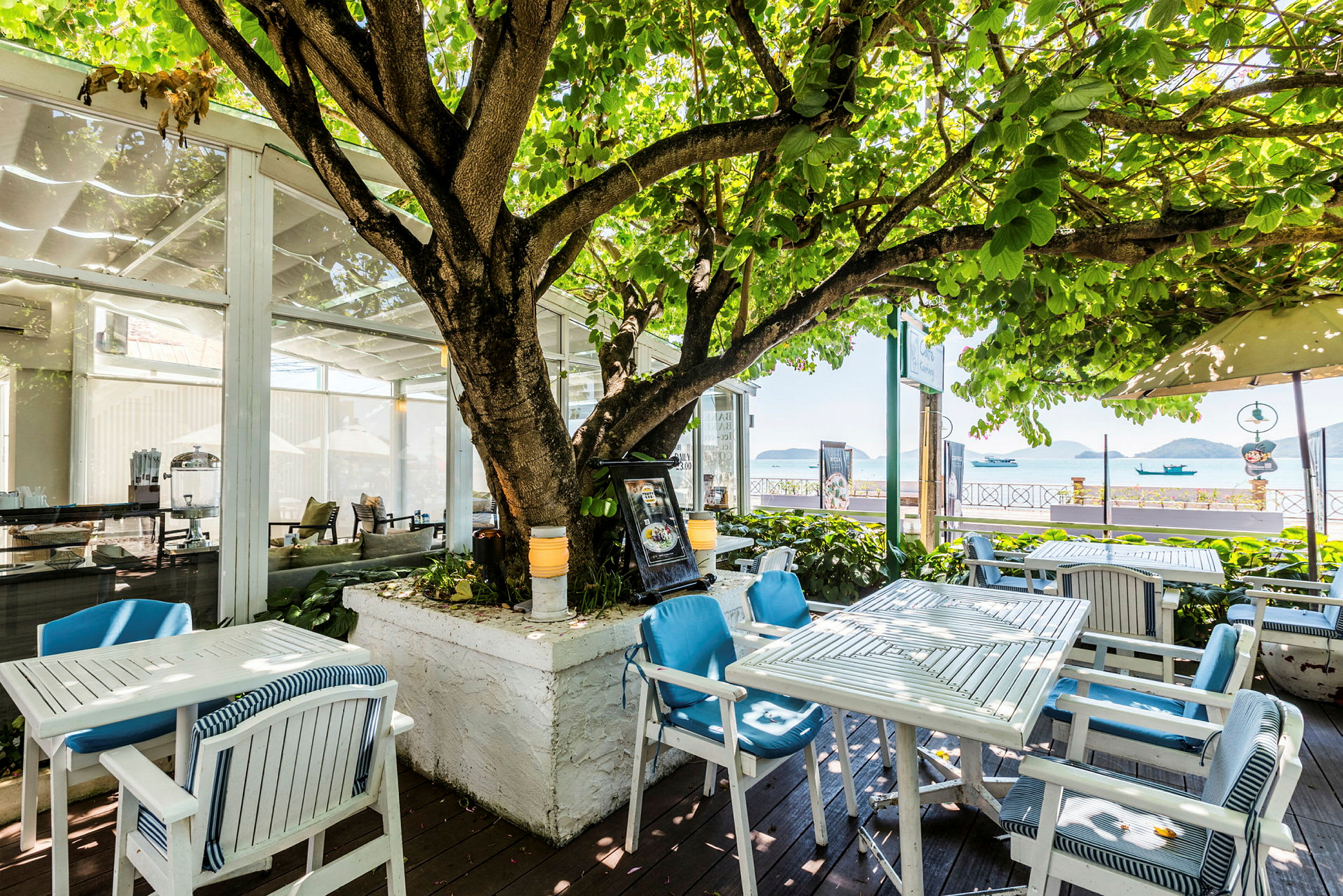 Utomhusrestaurang med bord och stolar under ett stort träd vid havet. (AI-genererad text)