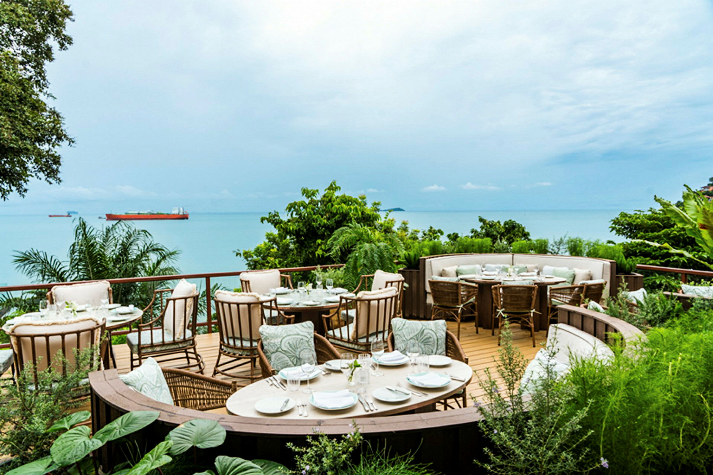 En utomhusrestaurang belägen på en terrass med utsikt över havet och frodig grönska. (AI-genererad text)