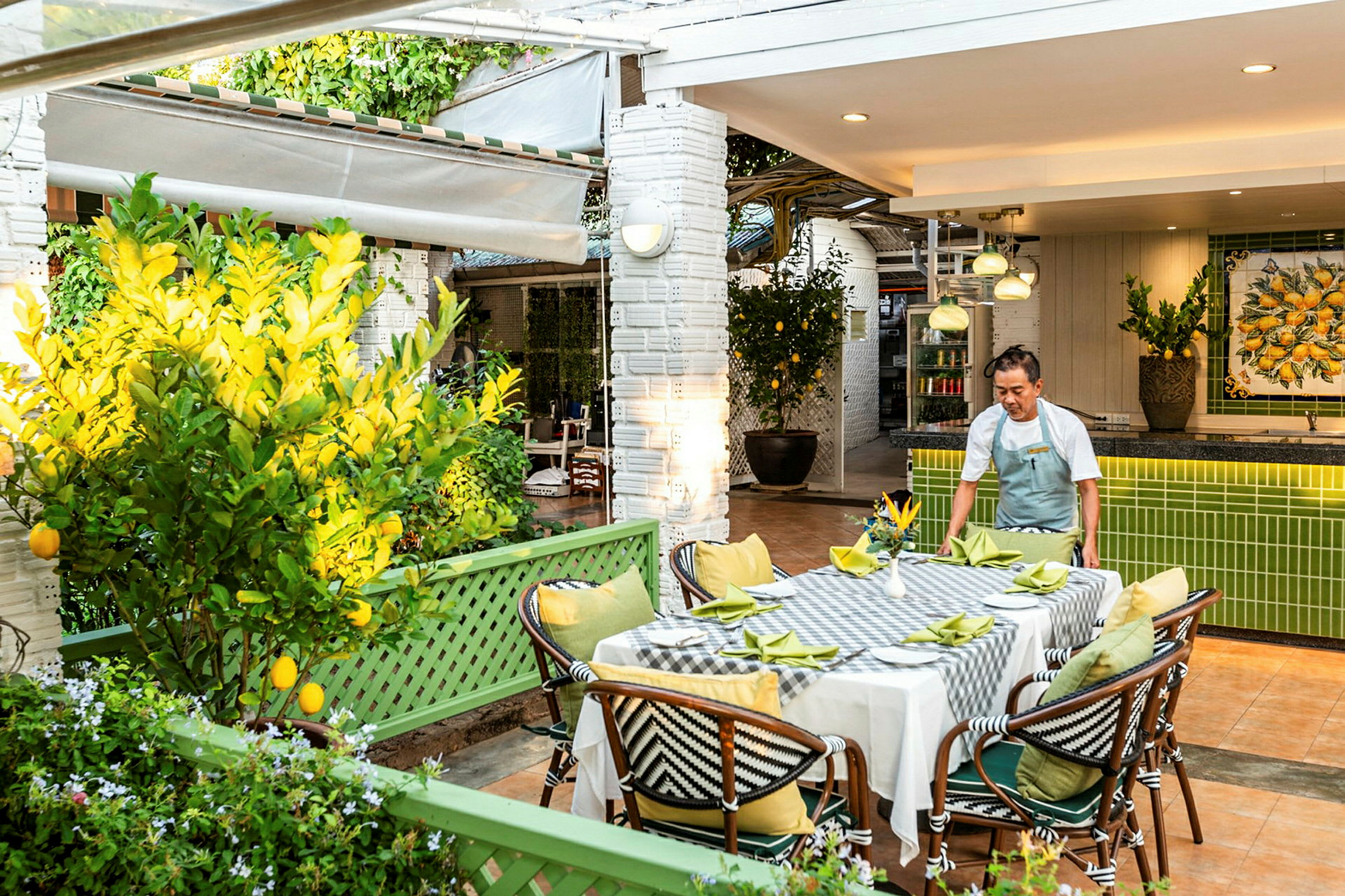 En utomhusrestaurang med bord klätt i vit duk och gröna servetter. En person arrangerar bordet. Området är omgivet av citrusträd och gröna växter. (AI-genererad text)