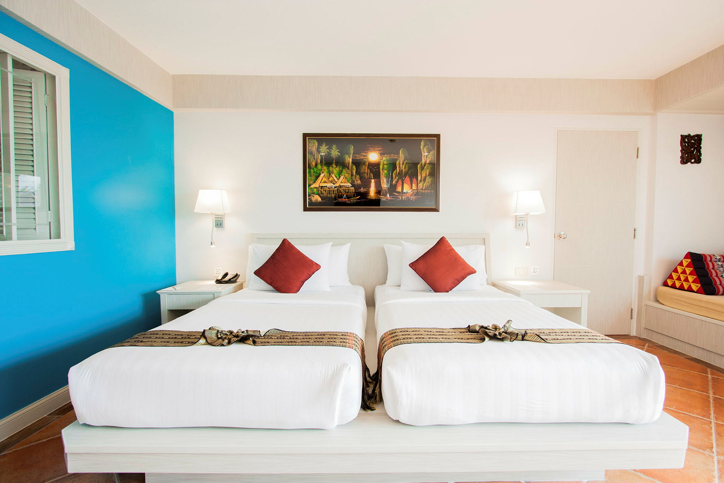 Ett hotellrum med två enkelsängar, vita sängkläder och dekorativa röda kuddar. På väggen hänger en färgglad tavla. (AI-genererad text)
