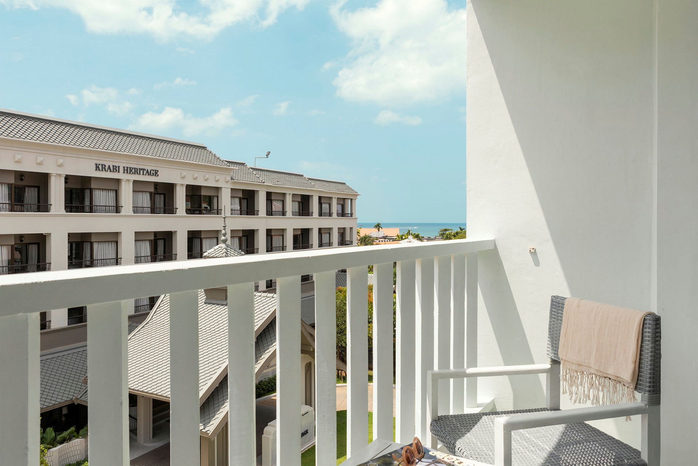 En balkong med utsikt över ett hotell med en skylt som lyder 'Krabi Heritage'. I bakgrunden syns havet och en klar himmel. (AI-genererad text)
