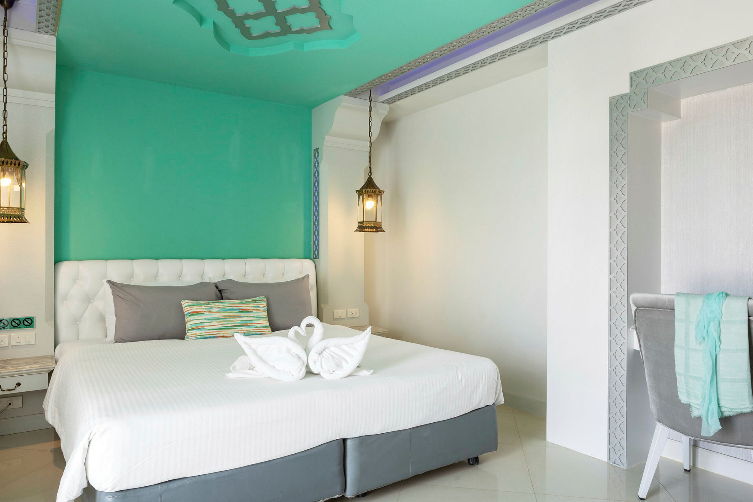 Ett elegant hotellrum med en vit dubbelsäng och gröna väggar. På sängen finns handdukar vikta till svanform och en färgglad kudde. Två hängande lampor skapar en behaglig atmosfär. (AI-genererad text)
