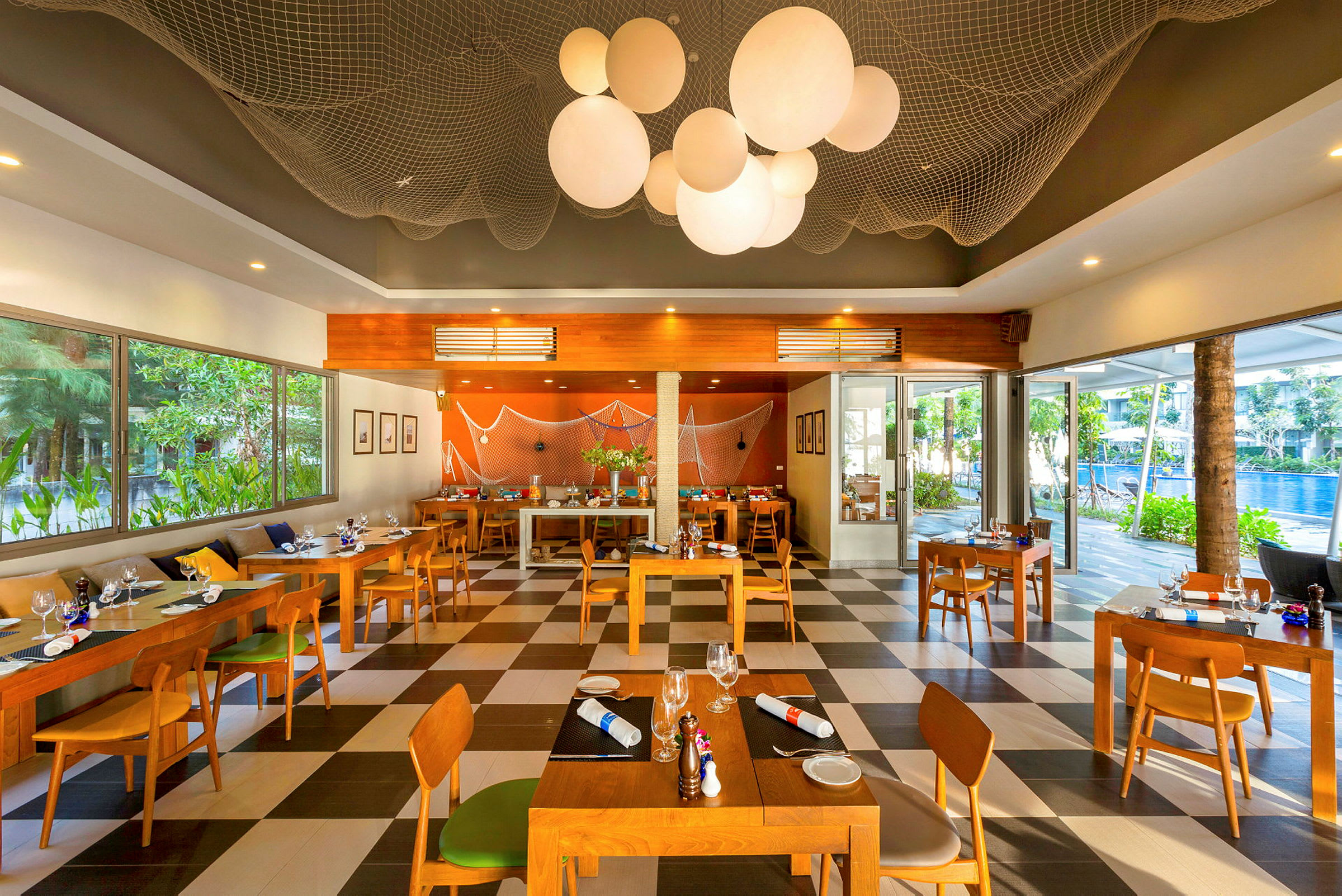 Restaurang med träbord och stolar samt stora fönster med utsikt över grönska och en pool utanför. (AI-genererad text)