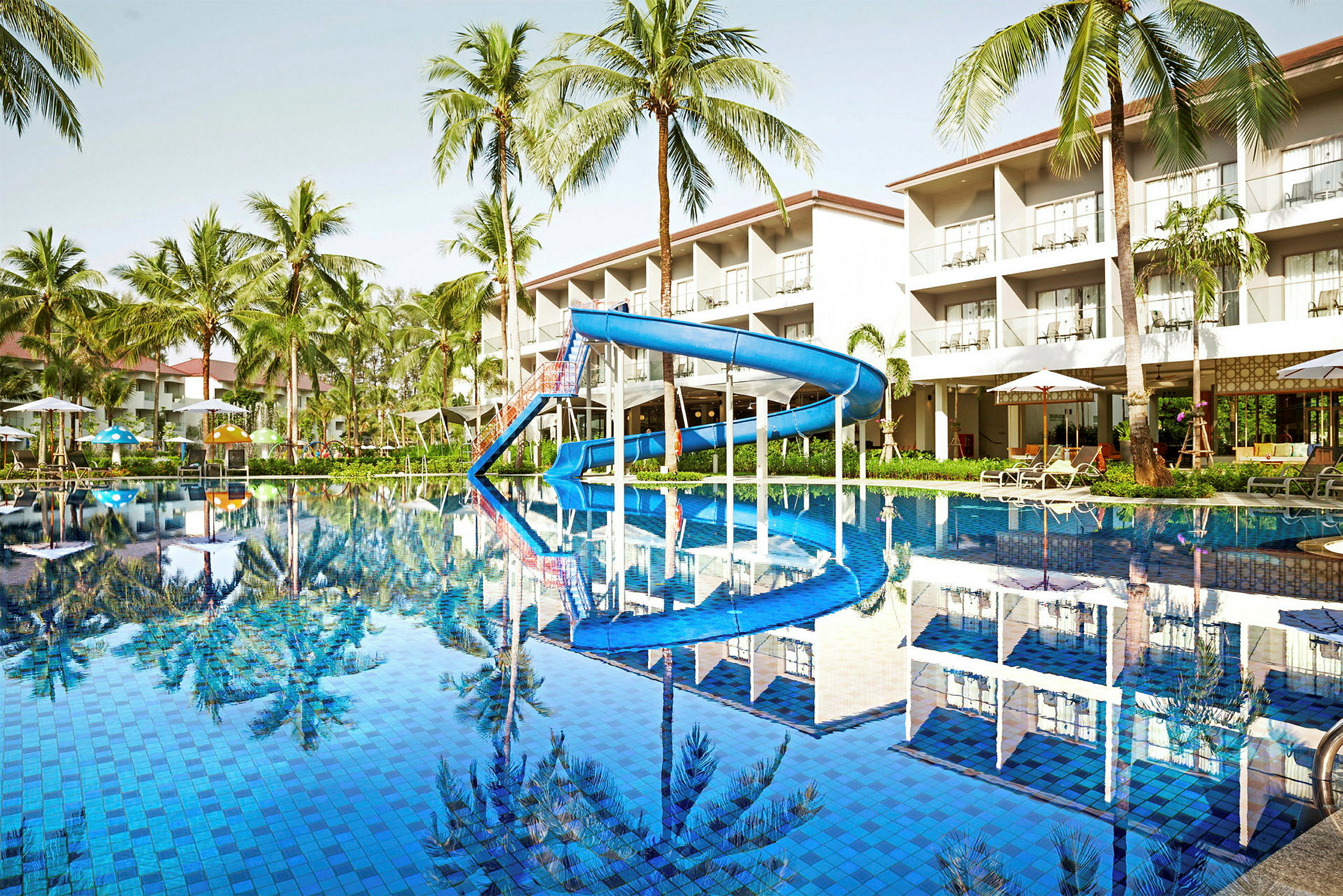 Bild av en pool med en blå vattenrutschkana, omgiven av solstolar och parasoller. Bakgrunden visar palmträd och en hotellbyggnad. (AI-genererad text)