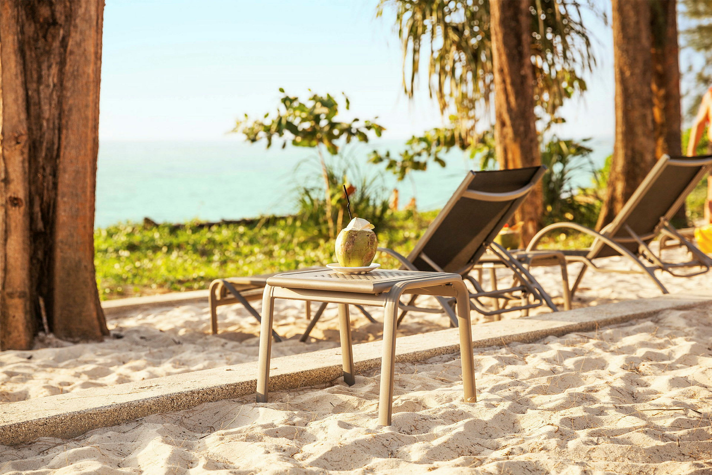 En kokosnöt med sugrör står på ett bord vid solstolar på stranden. (AI-genererad text)