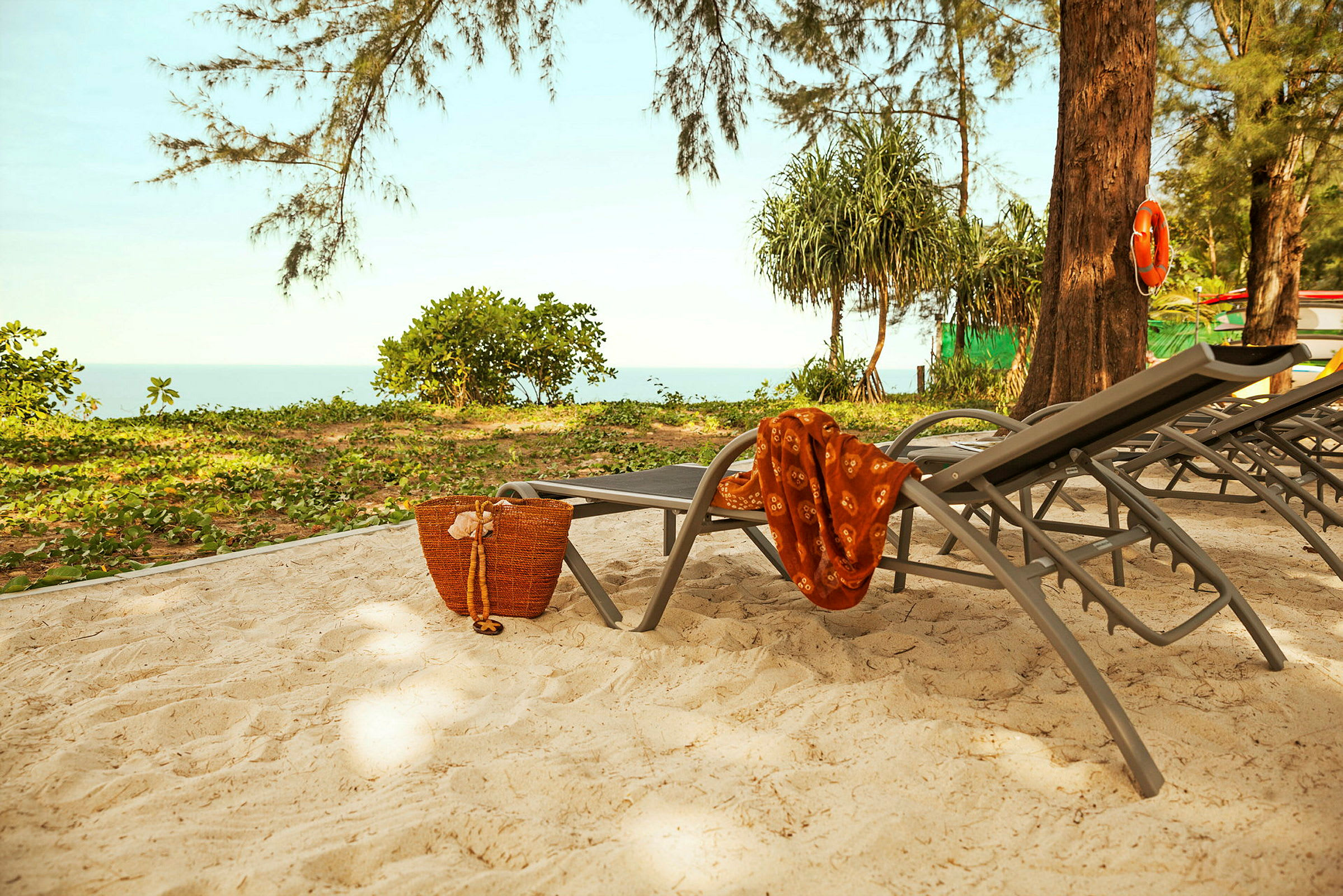 Solstolar på en sandig strand med grönska i bakgrunden, en korg och ett orange tyg ligger på en av stolarna. (AI-genererad text)