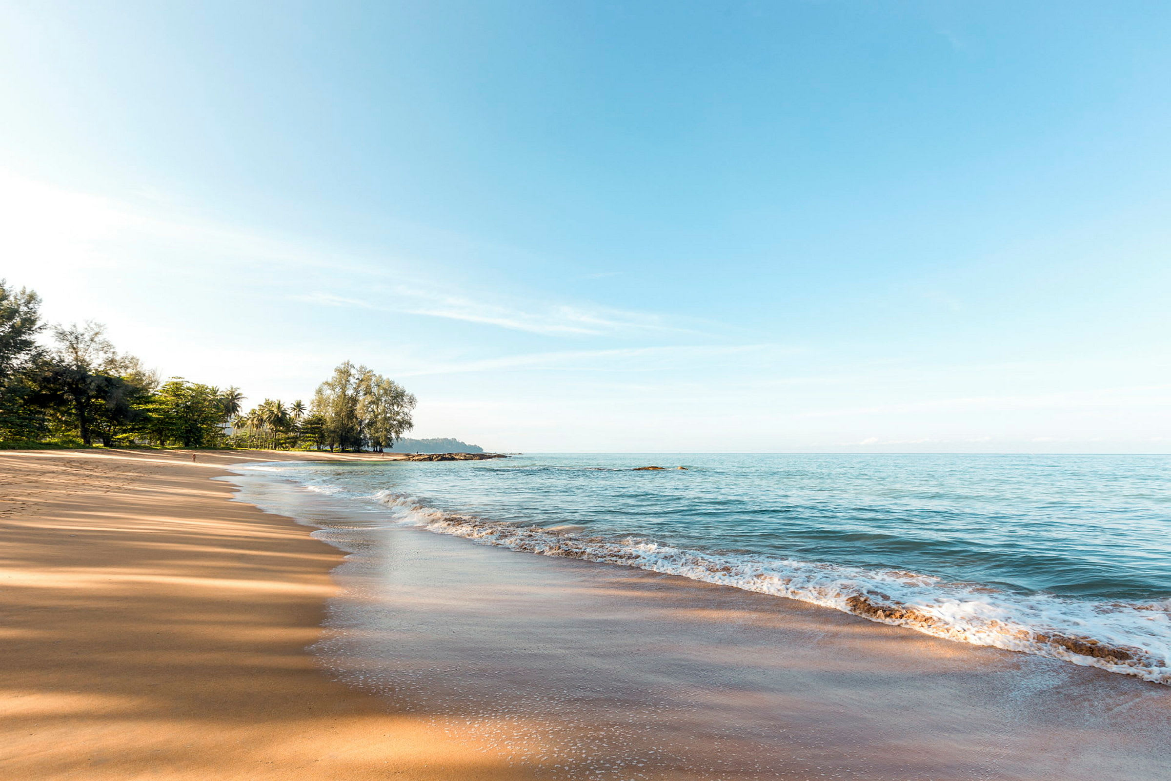 En sandstrand med havet och strandkant i förgrunden, träd syns i bakgrunden. (AI-genererad text)