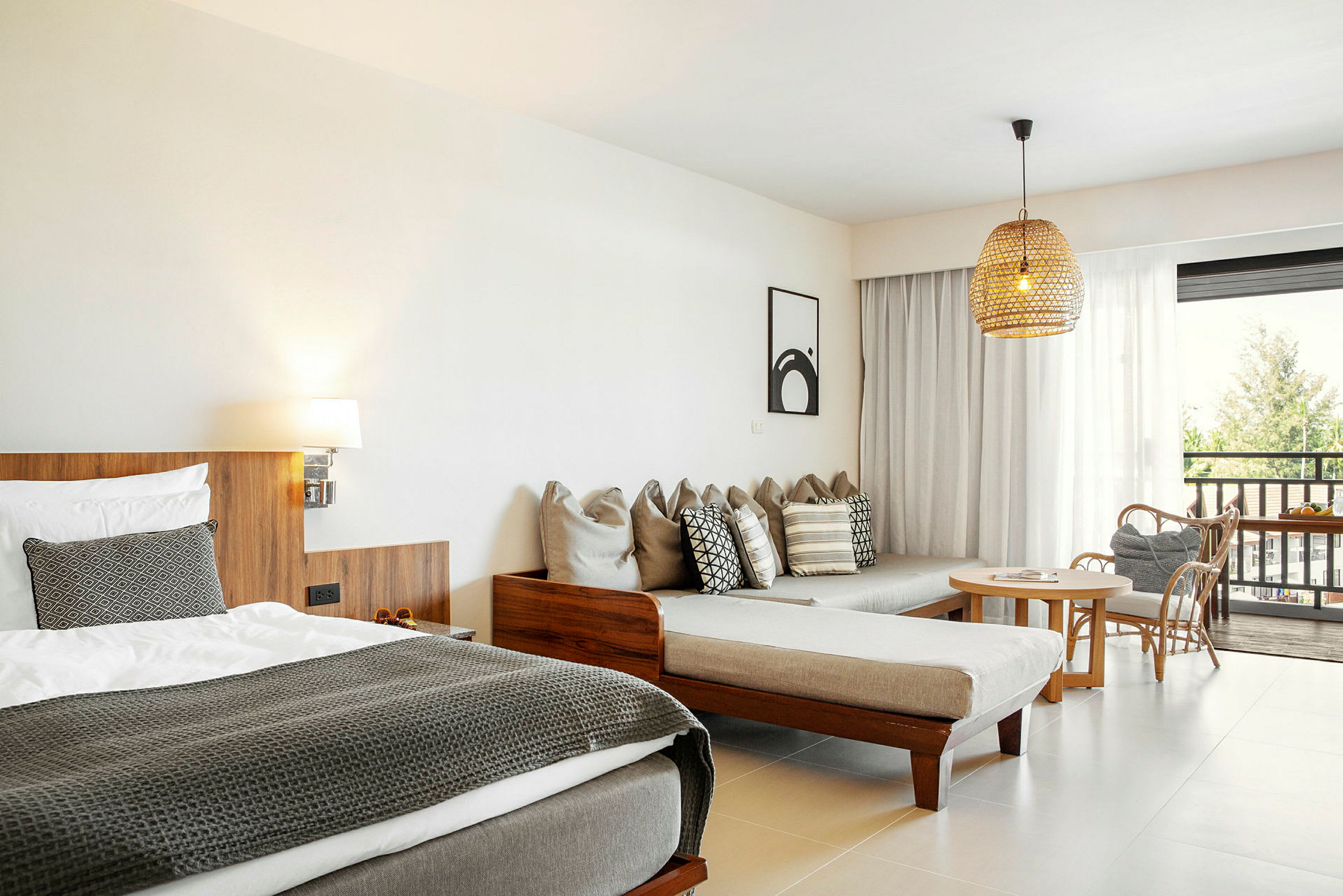 Ett hotellrum med en säng, en soffa med kuddar, ett bord med stolar och en utomhusutsikt genom glasdörrar. (AI-genererad text)