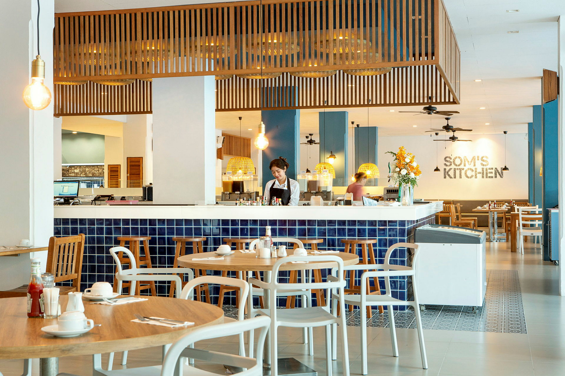 Bild av en restaurang med modern inredning och blåa kakel, där träbord och stolar är arrangerade runt omkring. (AI-genererad text)