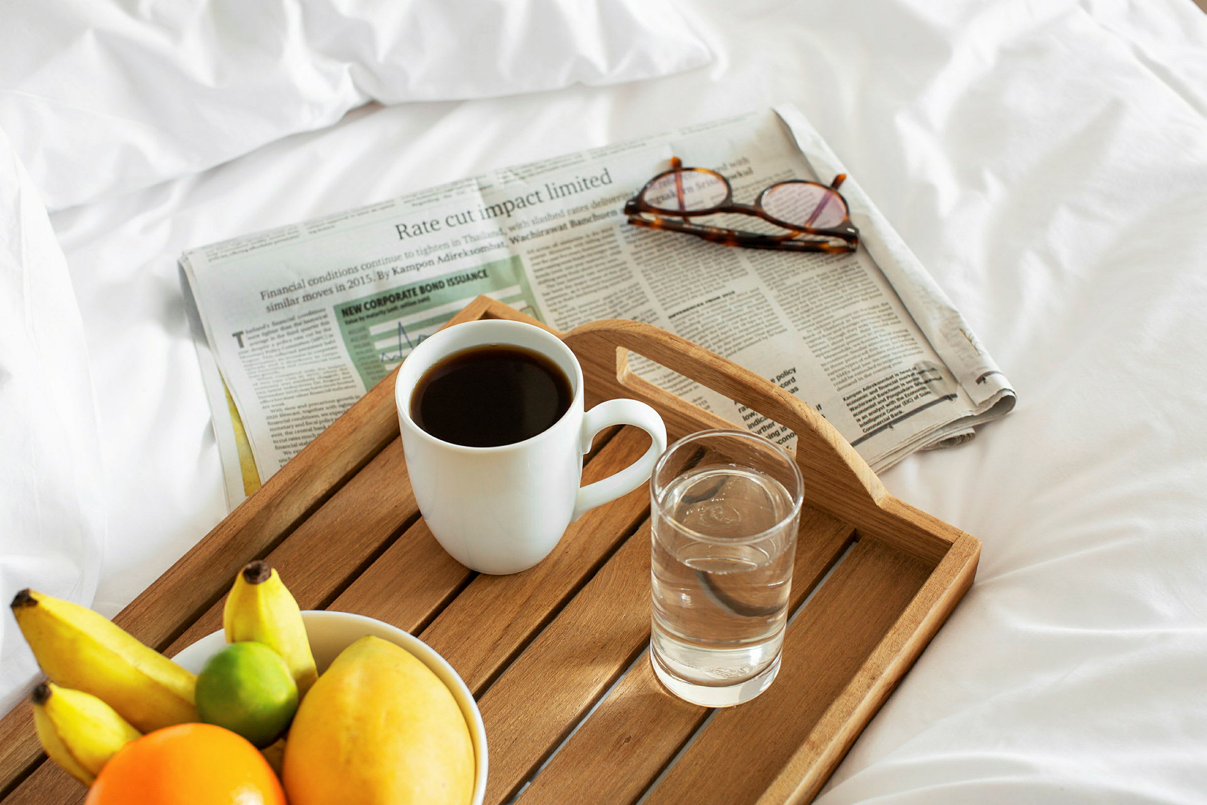 En frukostbricka med en kopp kaffe, ett glas vatten, och en skål med frukt på en säng, bredvid en tidning och ett par glasögon. (AI-genererad text)