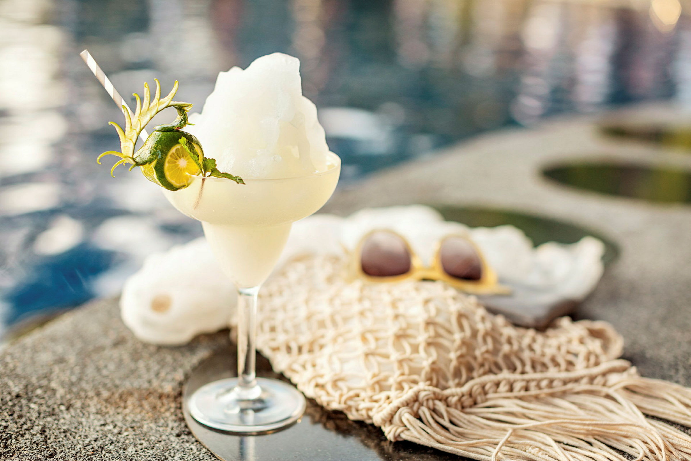 Ett glas fylld med en isig cocktail dekorerad med lime och mynta står på kanten bredvid en pool. (AI-genererad text)