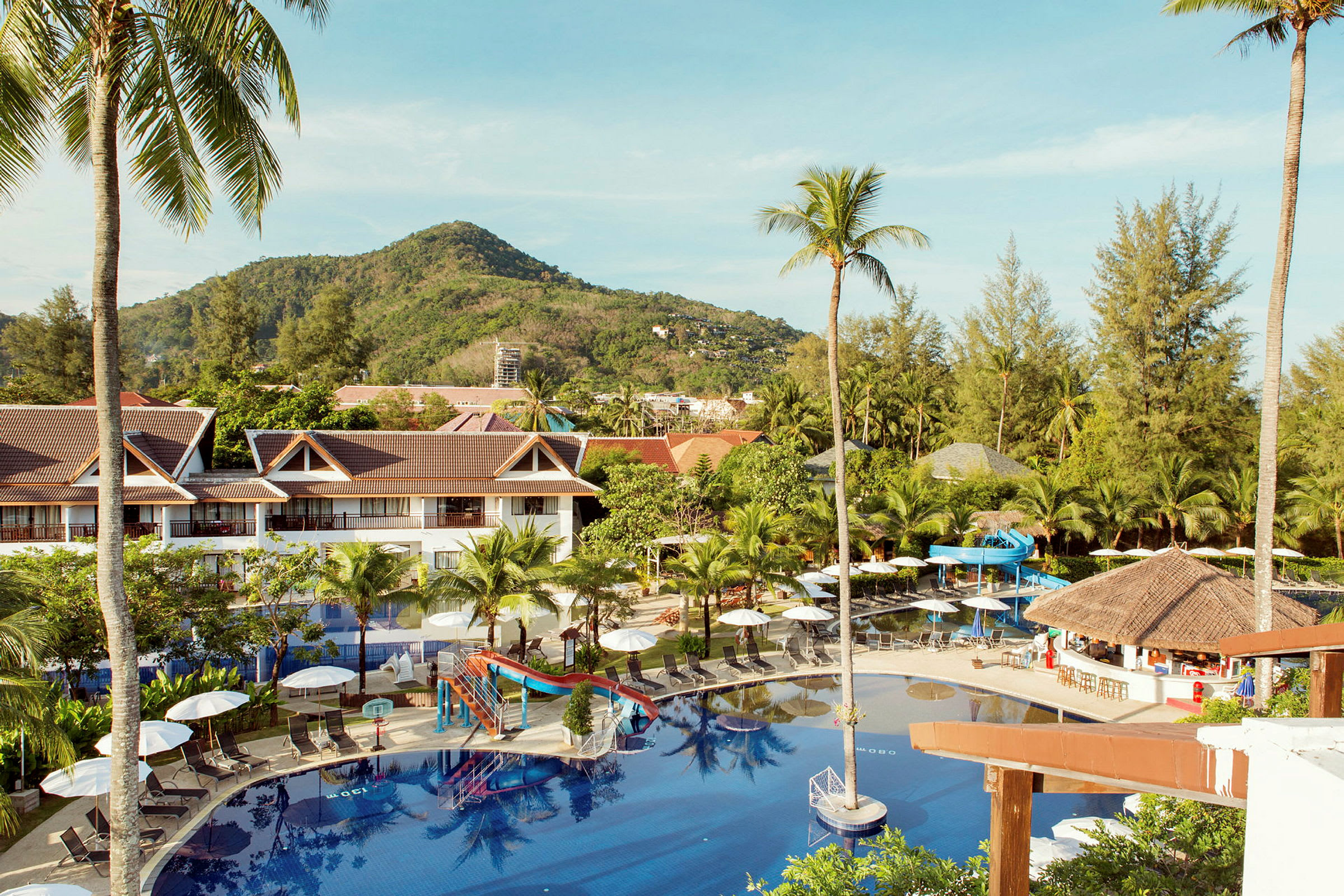 Utsikt över hotellområde med pool, solsängar och parasoller, omgiven av grönska och berg. (AI-genererad text)