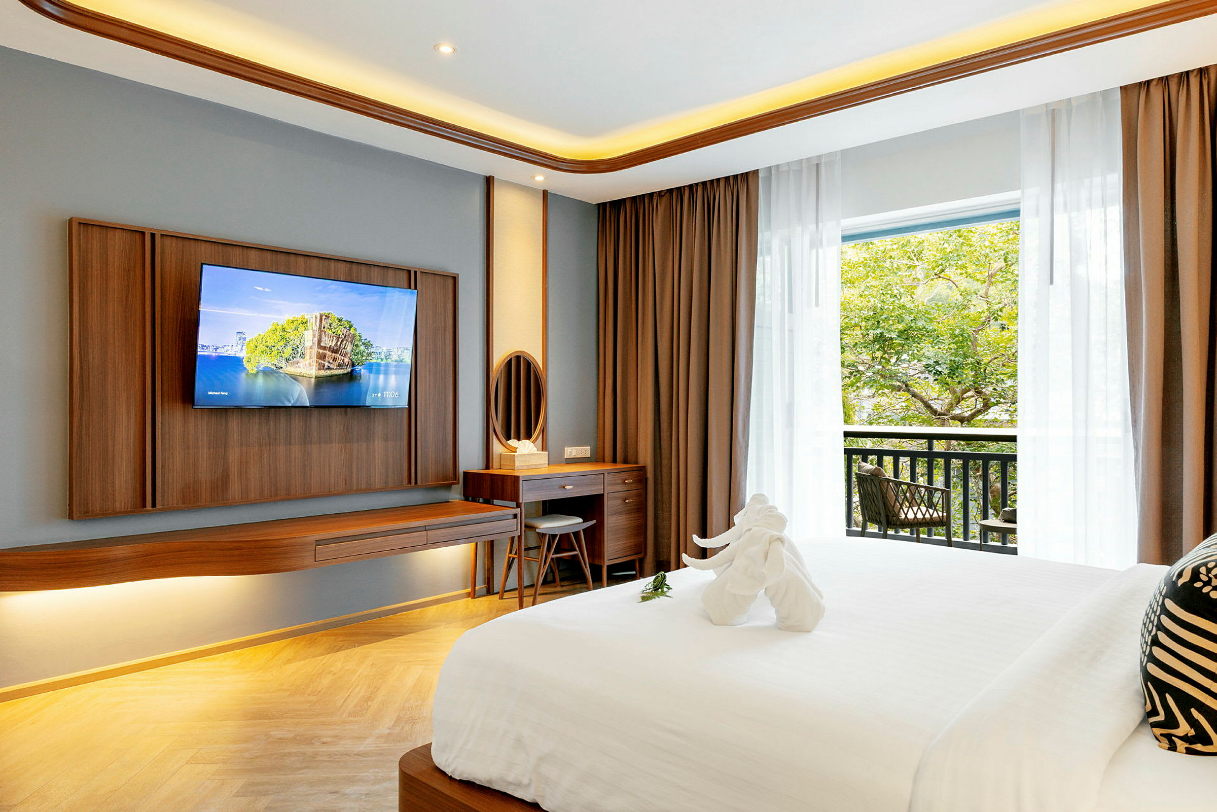 Ett hotellrum med modern inredning inklusive en stor säng, en väggmonterad tv, ett skrivbord med en spegel och en balkong med utsikt över grönska. (AI-genererad text)
