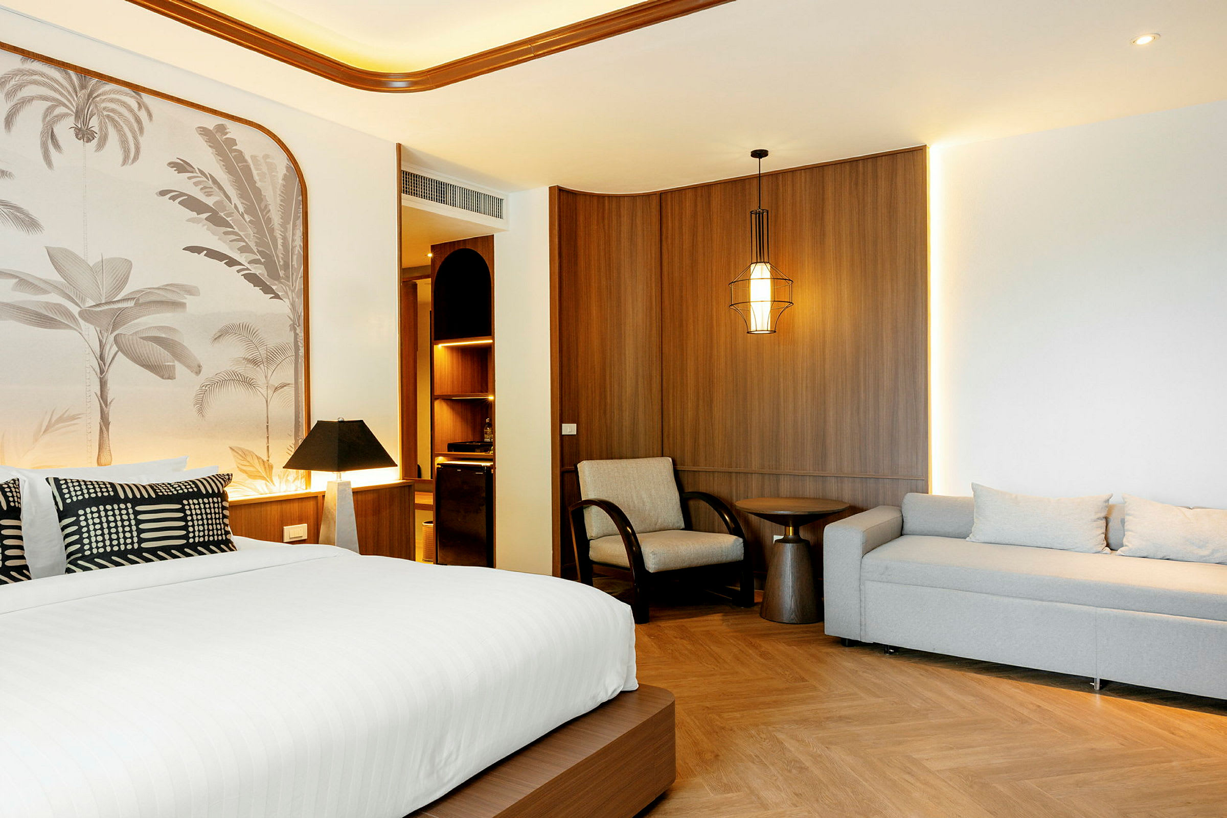 Ett hotellrum med en stor säng, vita sängkläder, en grå soffa, en fåtölj och ett soffbord. Väggarna har träpanel och en dekorativ tapet. (AI-genererad text)